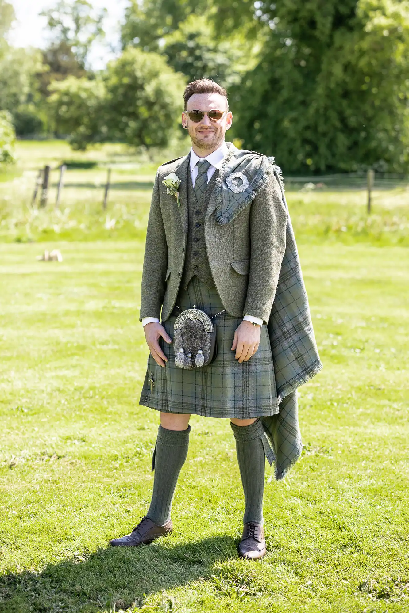 A man in sunglasses is outdoors wearing a green tartan kilt ensemble with a jacket, vest, sporran, and traditional Scottish accessories. He is standing on a grassy area with trees in the background.