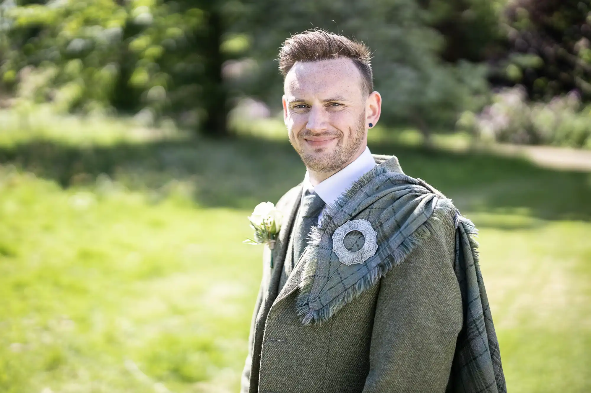 A man in traditional Scottish attire, featuring a green jacket and tartan sash, stands outdoors on grass with trees in the background.