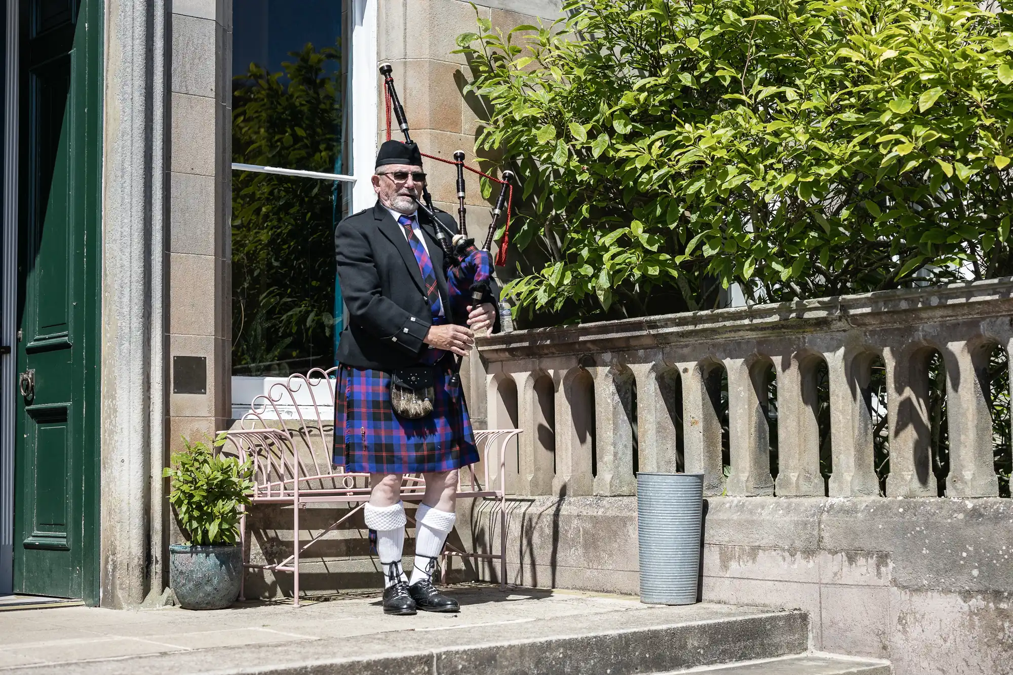 A man in traditional Scottish attire plays the bagpipes outside next to a stone railing and potted plants.