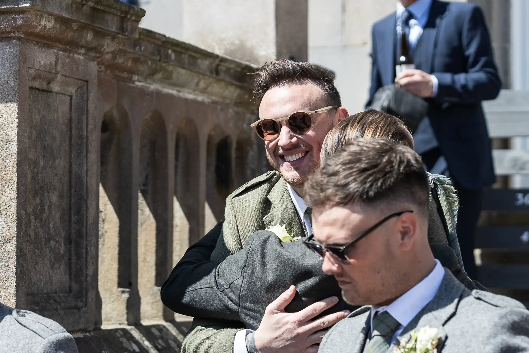 A man in sunglasses hugs someone while another man in a suit and tie sits nearby. They are outdoors, possibly at a formal event.