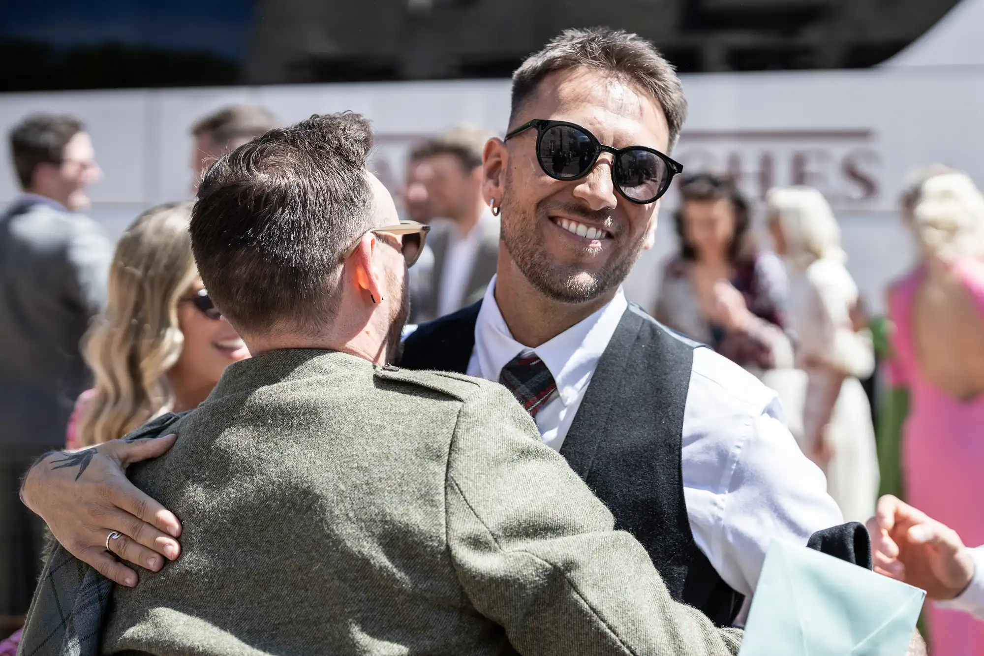 Two men hugging at a social event, one in sunglasses and a vest smiling. A woman in sunglasses is partially visible in the background. Other people in formal attire are seen in the distance.