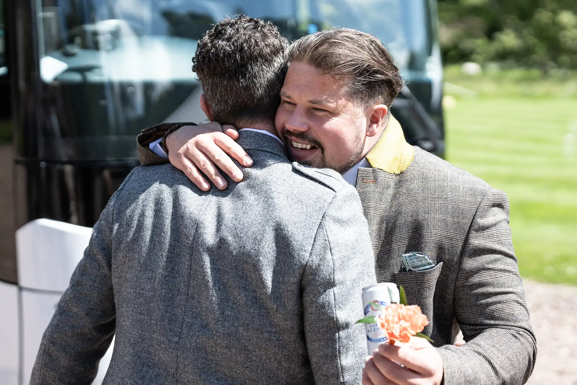 Two men in suits embrace outdoors near a bus. One man holds a drink can and a flower.