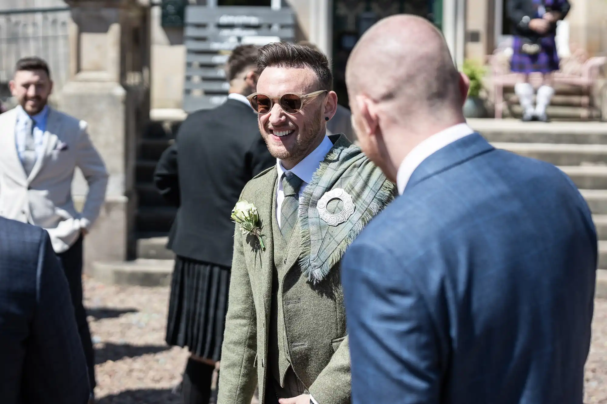 A man in a green suit and sunglasses stands smiling and talking with another man in a blue suit at an outdoor gathering. Other people are visible in the background.