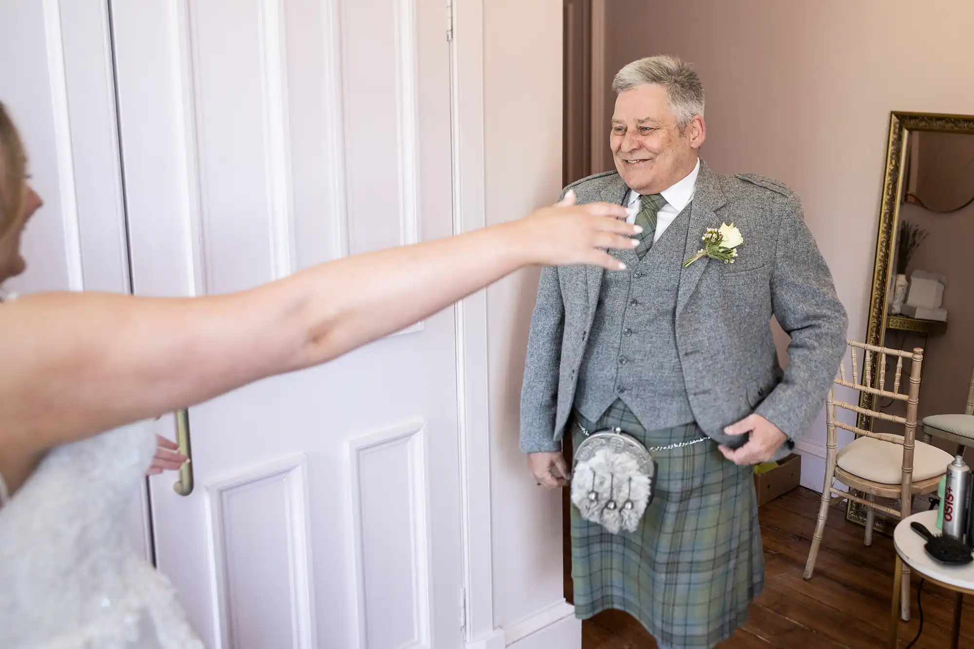 A man in a gray kilt suit smiles as he is greeted by a woman reaching towards him with open arms in a room.