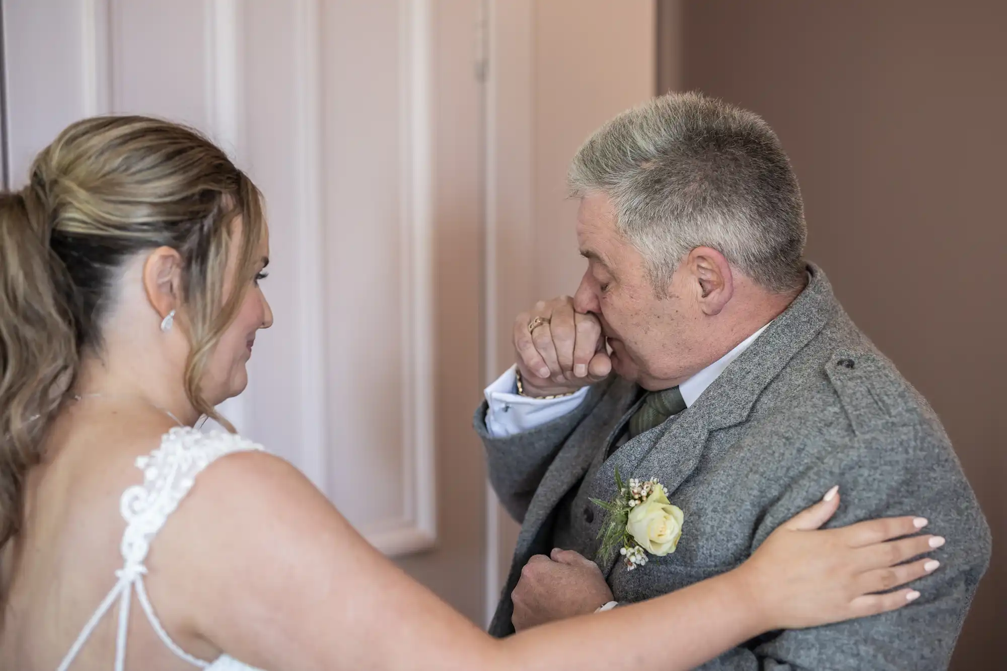 A woman in a bridal gown gently touches an older man's shoulder. The man, dressed in a gray suit with a boutonniere, appears emotional with his hand covering his mouth.