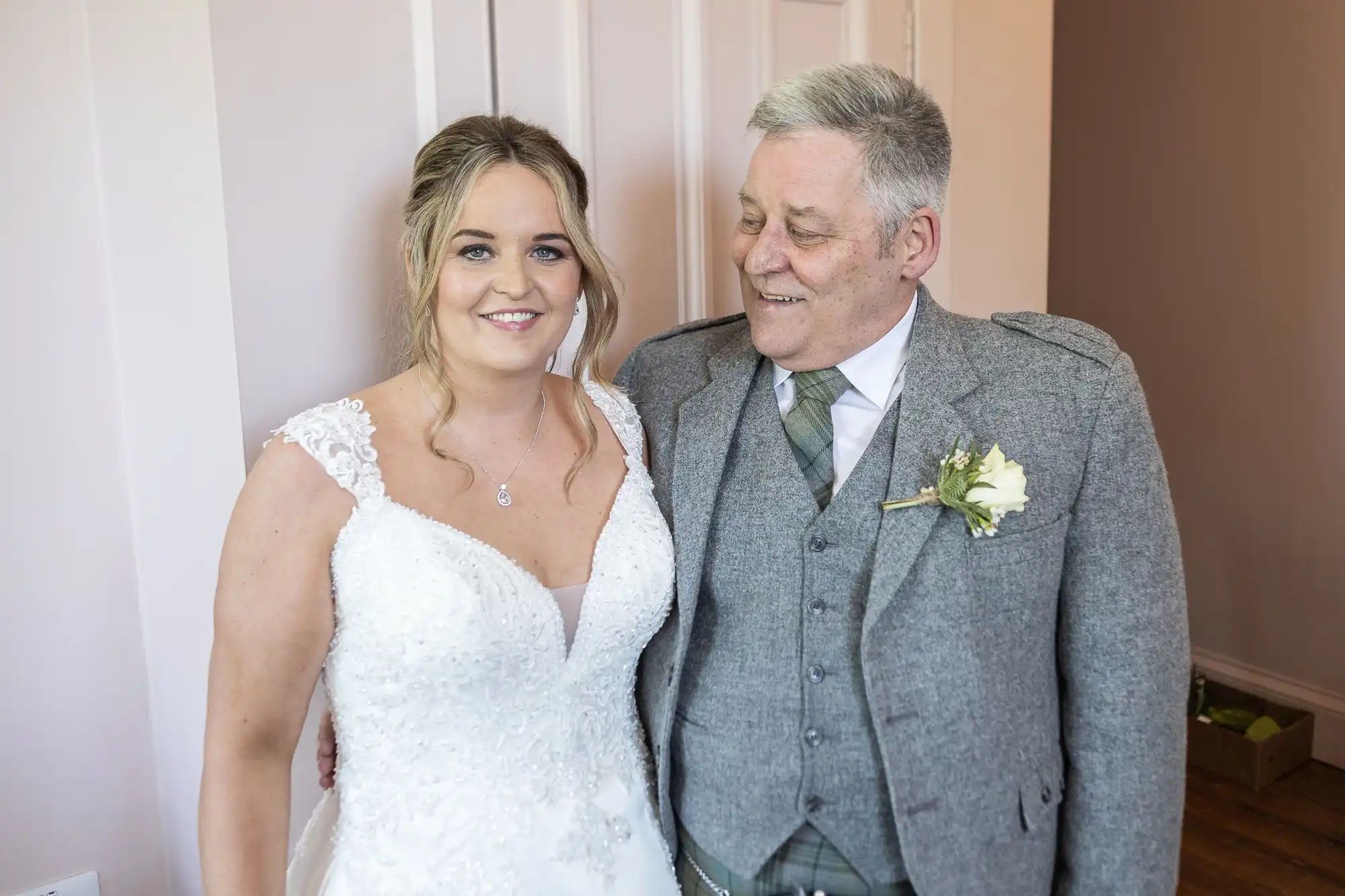 A woman in a white wedding dress stands beside an elderly man in a grey suit and tie. Both are smiling and standing in front of a light-colored wall indoors.