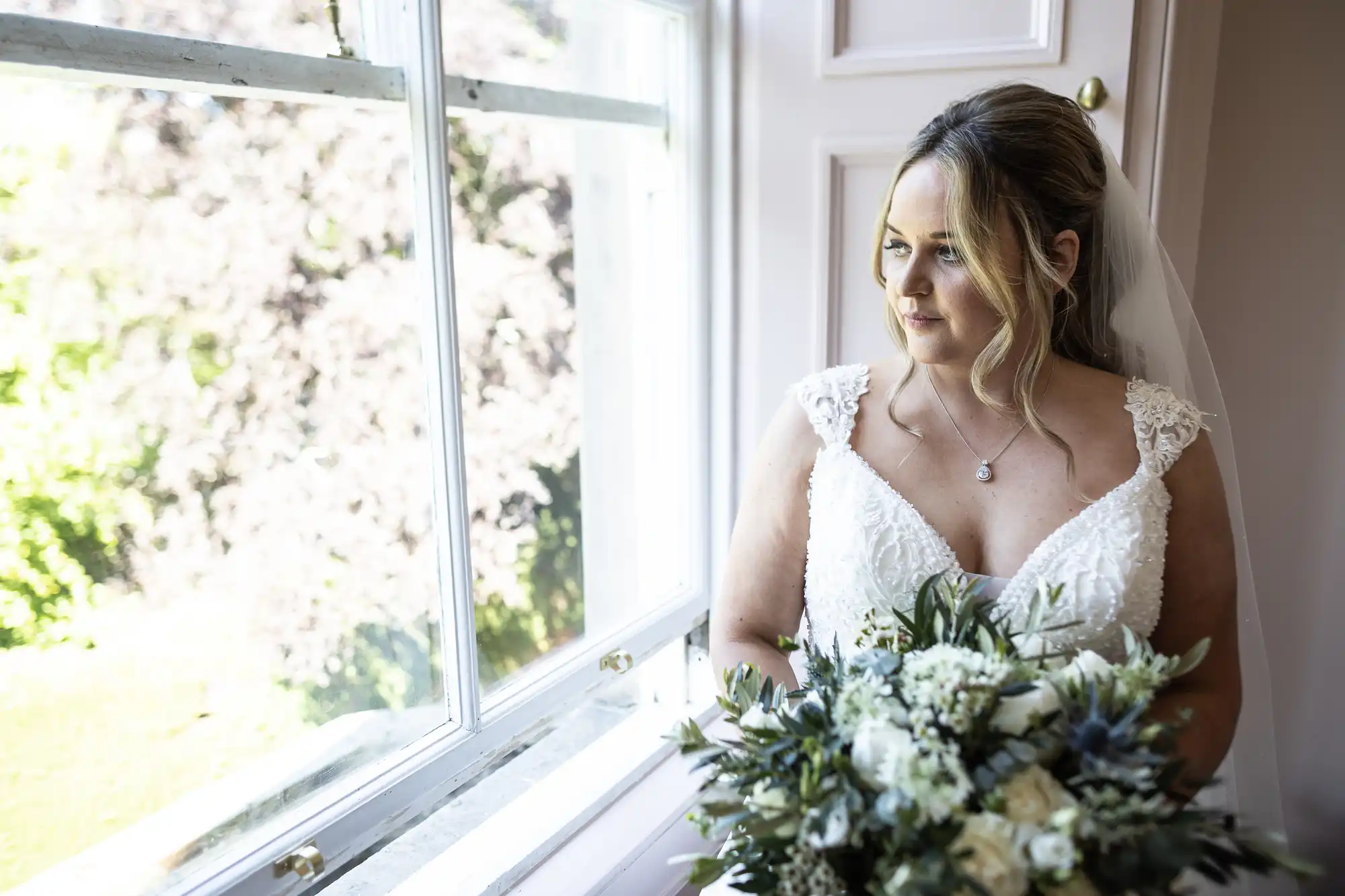 A bride in a white wedding dress stands by a window, holding a bouquet of flowers, and looks outside.