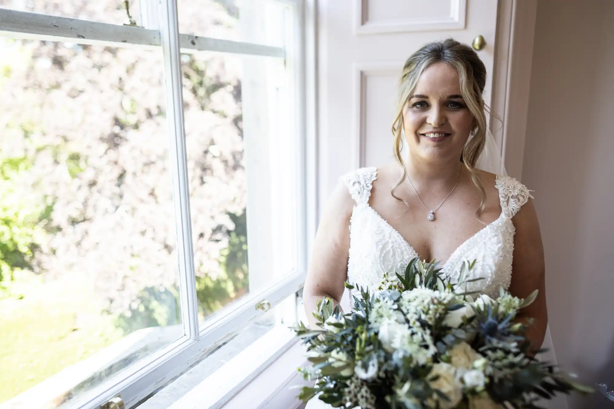 A woman in a white wedding dress stands next to a window, holding a bouquet of flowers, and smiling at the camera.