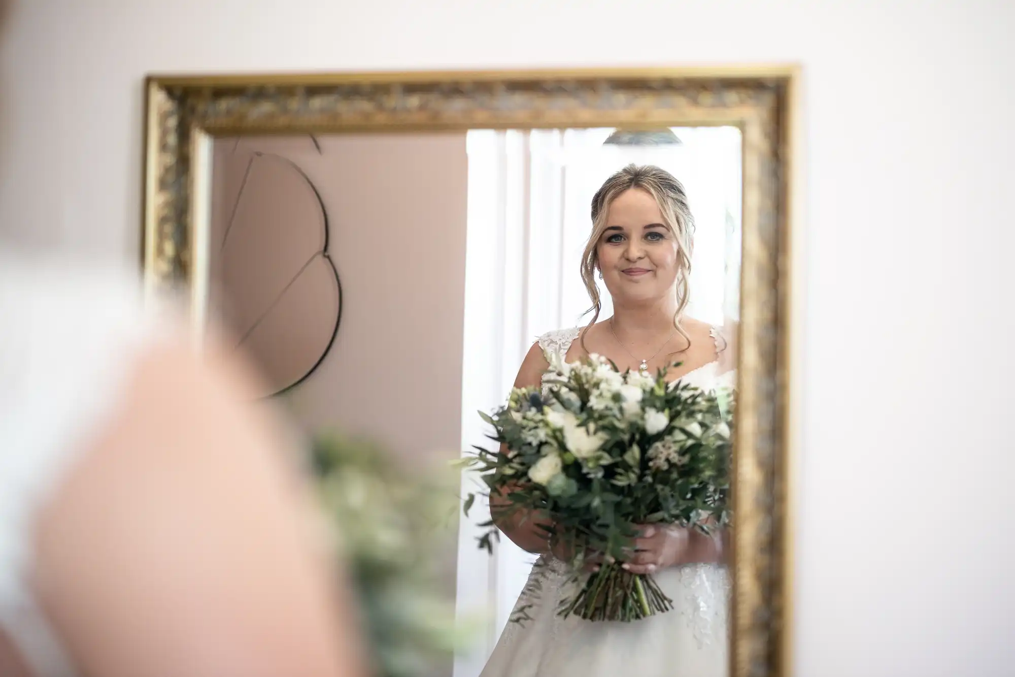 A bride holding a bouquet of flowers is reflected in a gold-framed mirror.