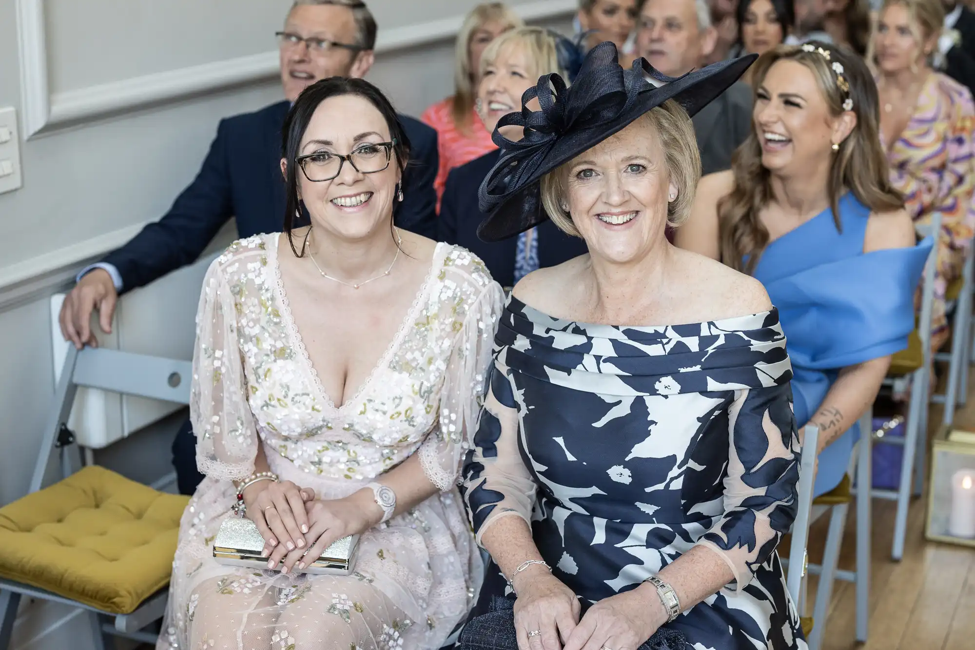 Two women in formal attire, one wearing a pale pink lace dress and glasses, the other in a black and white floral dress with a black hat, seated and smiling at a social event with others in the background.