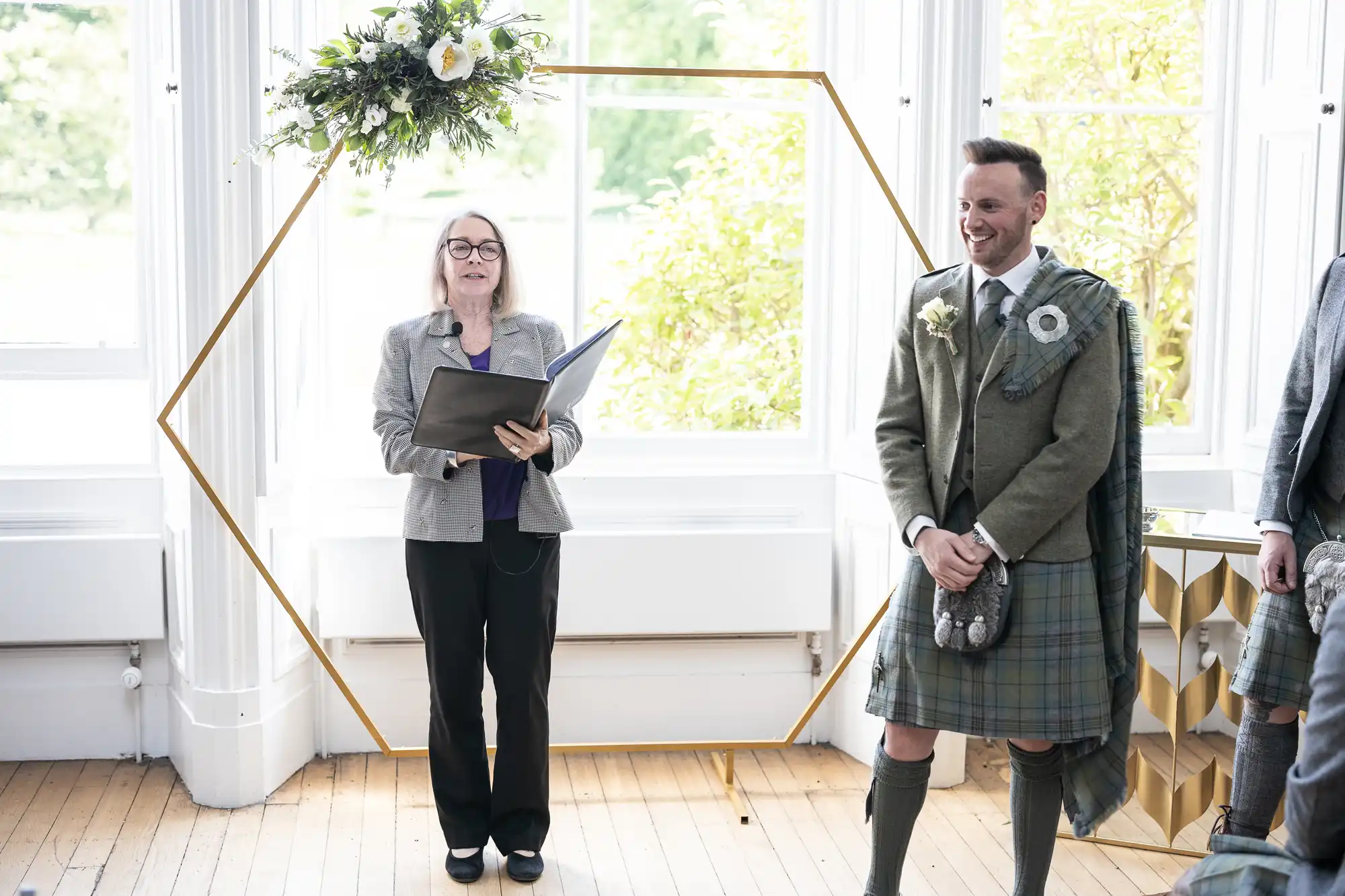 A person reads from a book while another person, dressed in traditional Scottish attire with a kilt and sporran, stands beside them in a bright room with large windows and a floral arrangement.