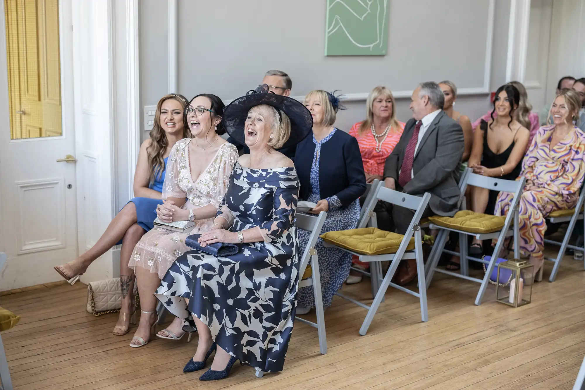 A group of well-dressed people sitting on folding chairs in a brightly lit room, some smiling and engaged in conversation, awaiting an event or ceremony.