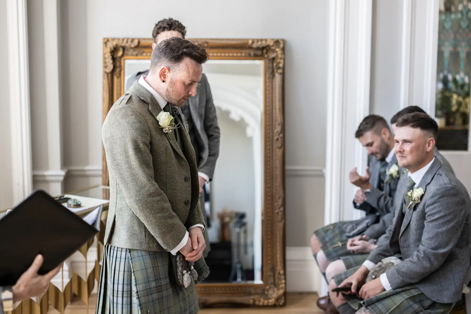 A groom in a kilt stands in thought next to a large mirror, while three groomsmen in kilts sit to the side, one holding a smartphone, in an elegantly furnished room.