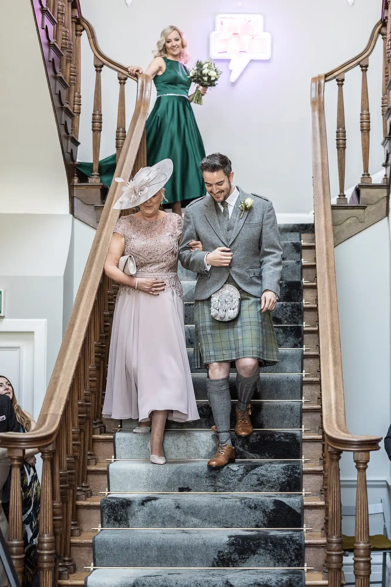 A man in a kilt escorts a woman in a pink dress and hat down a staircase at an event while another woman in a green dress descends behind them holding a bouquet.