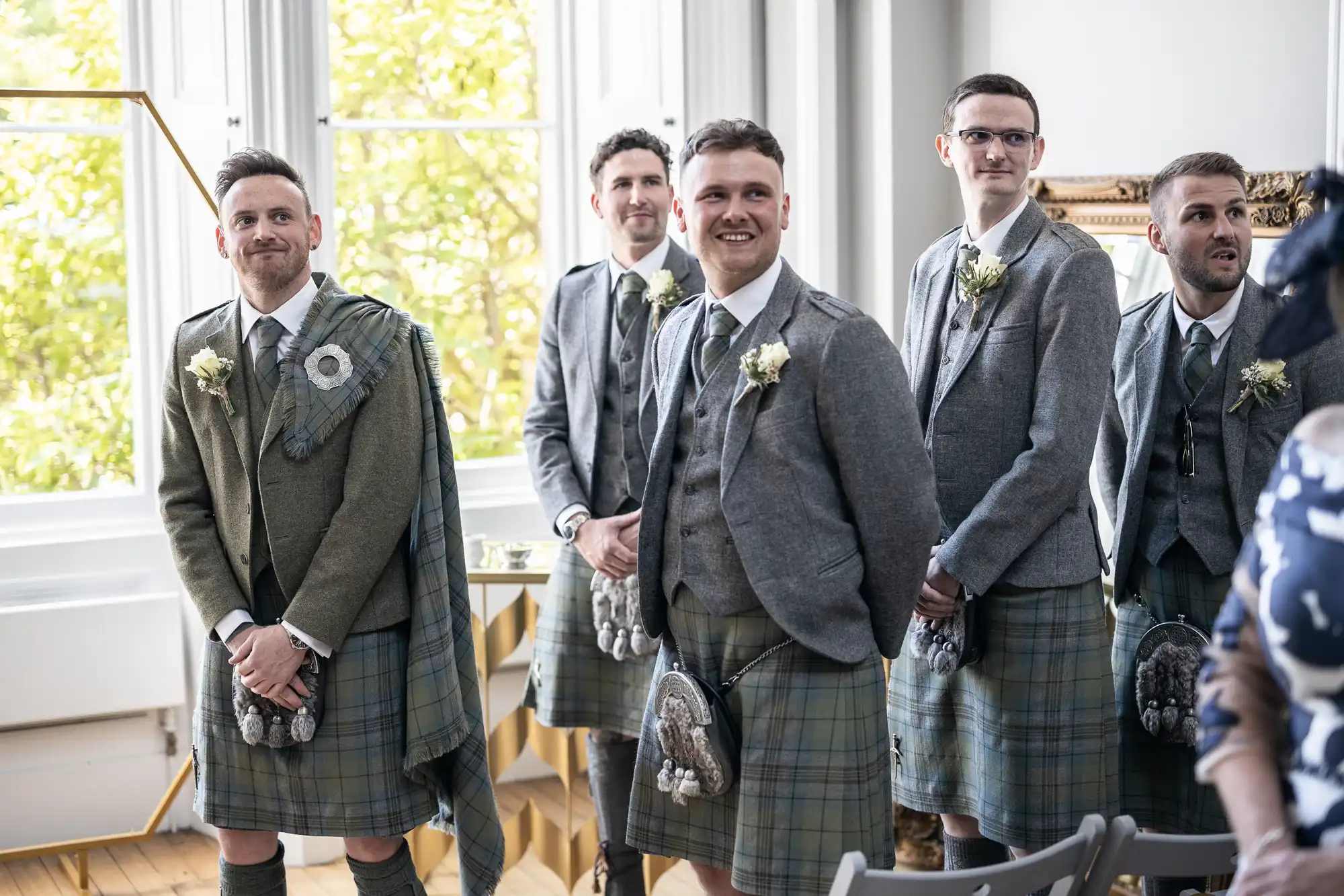 Five men wearing traditional Scottish kilts stand indoors smiling and looking towards the camera, with natural light coming through large windows.