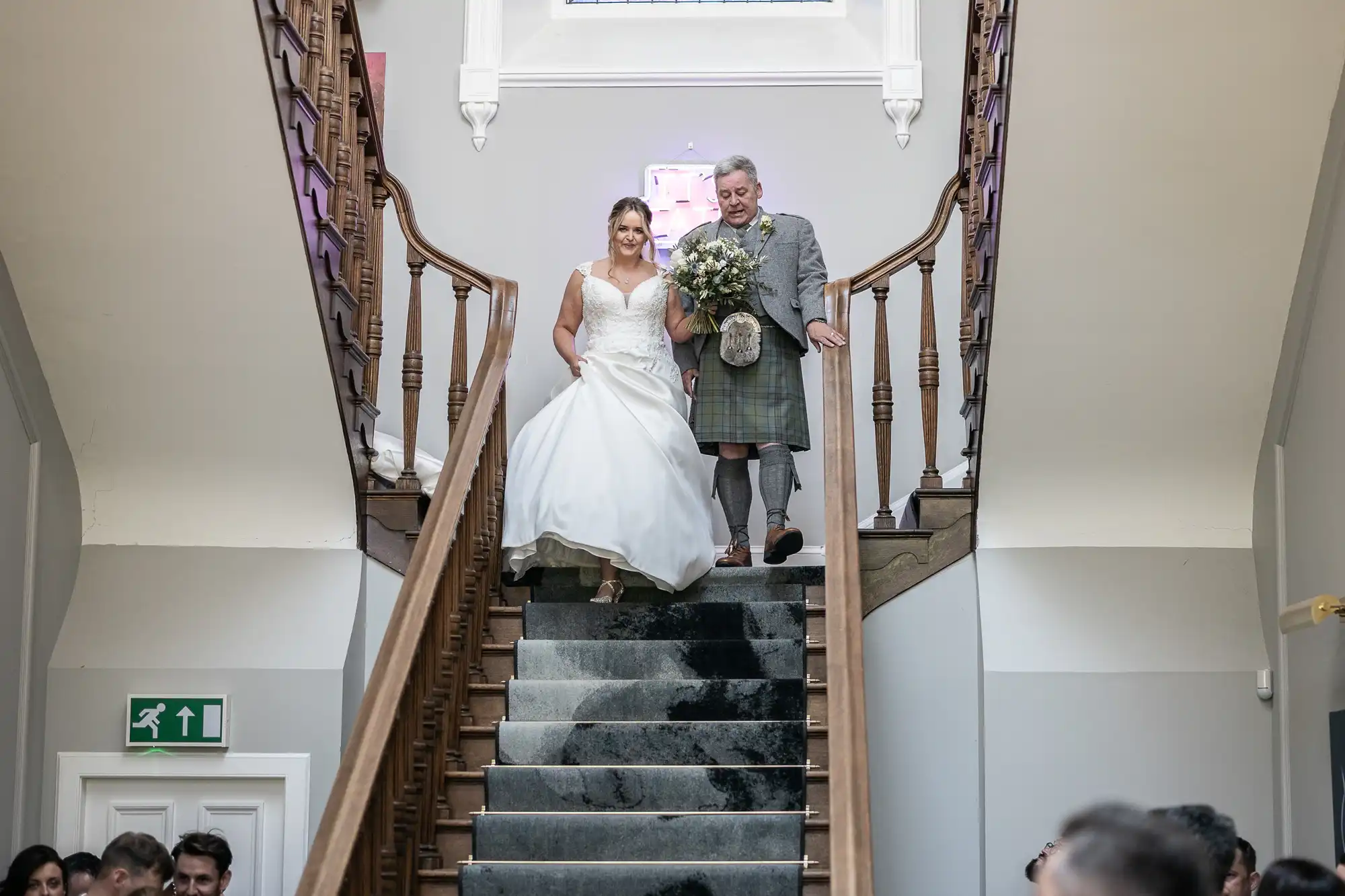 A bride in a white gown descends a staircase accompanied by an older man in a kilt, holding a bouquet. People are gathered at the bottom of the stairs.