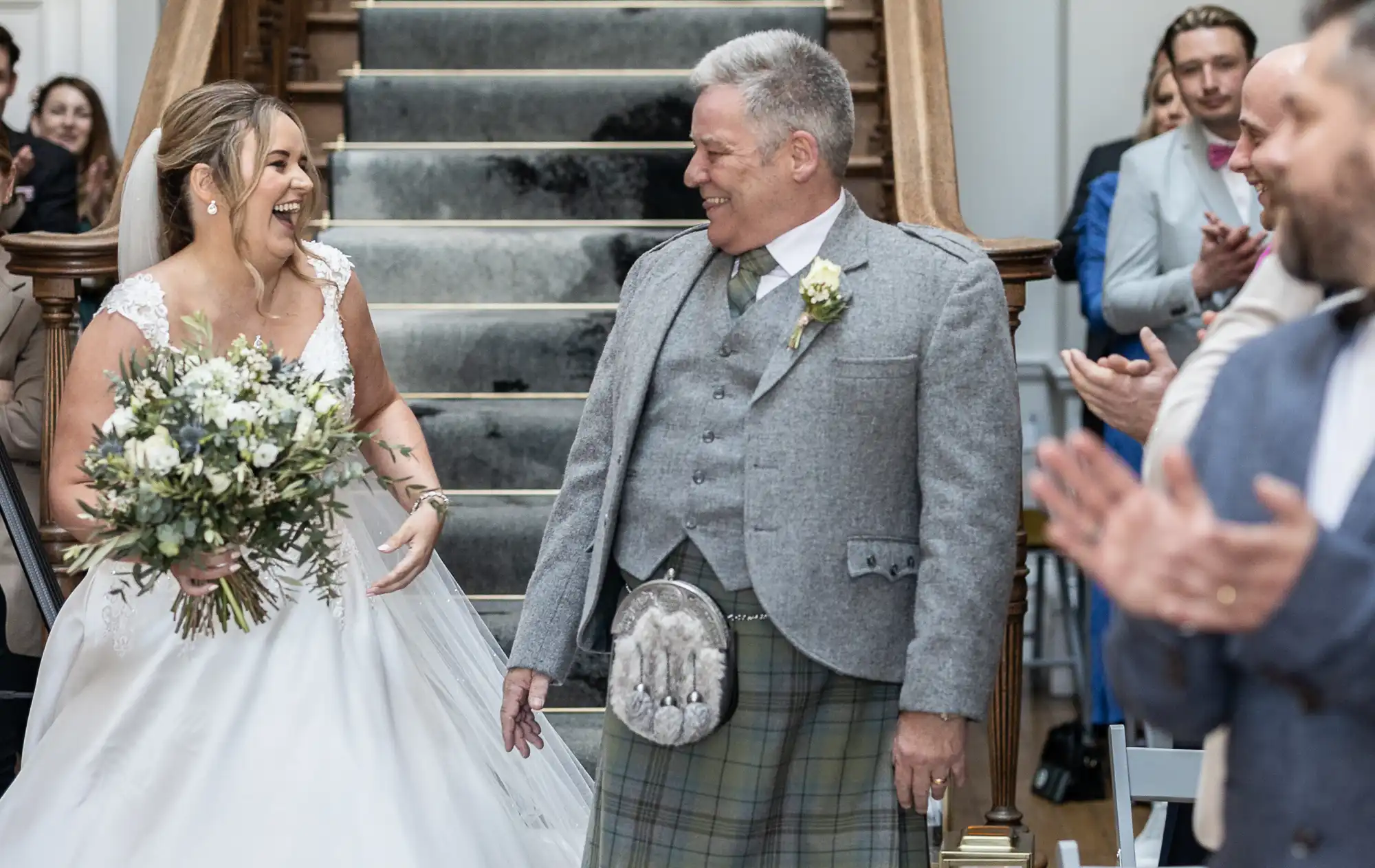 A bride in a white dress and a man in a gray suit with a kilt smile at each other while walking down a staircase, with onlookers clapping in the background.