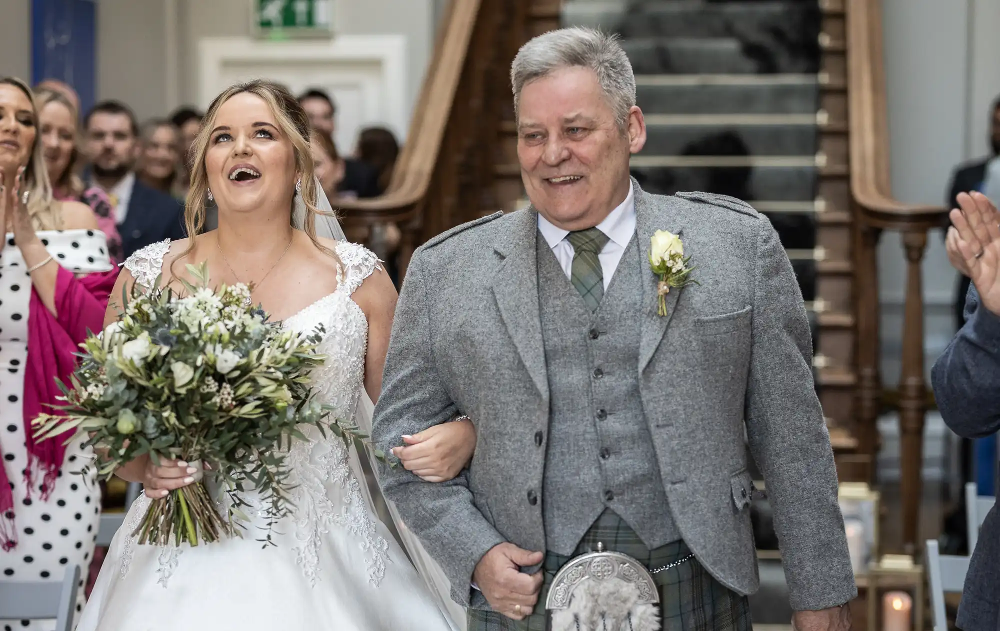 A bride in a white gown and a man in a gray suit walk arm-in-arm down the aisle, smiling, while guests applaud in the background.