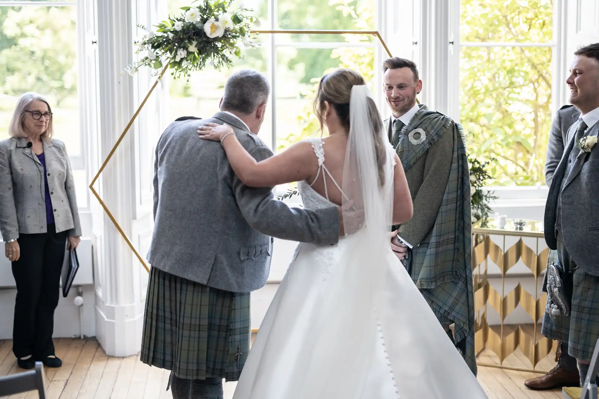 A bride, escorted by a man in a kilt, walks down the aisle towards a groom in a kilt. Two other people in kilts, one standing and one sitting, are visible in the background.