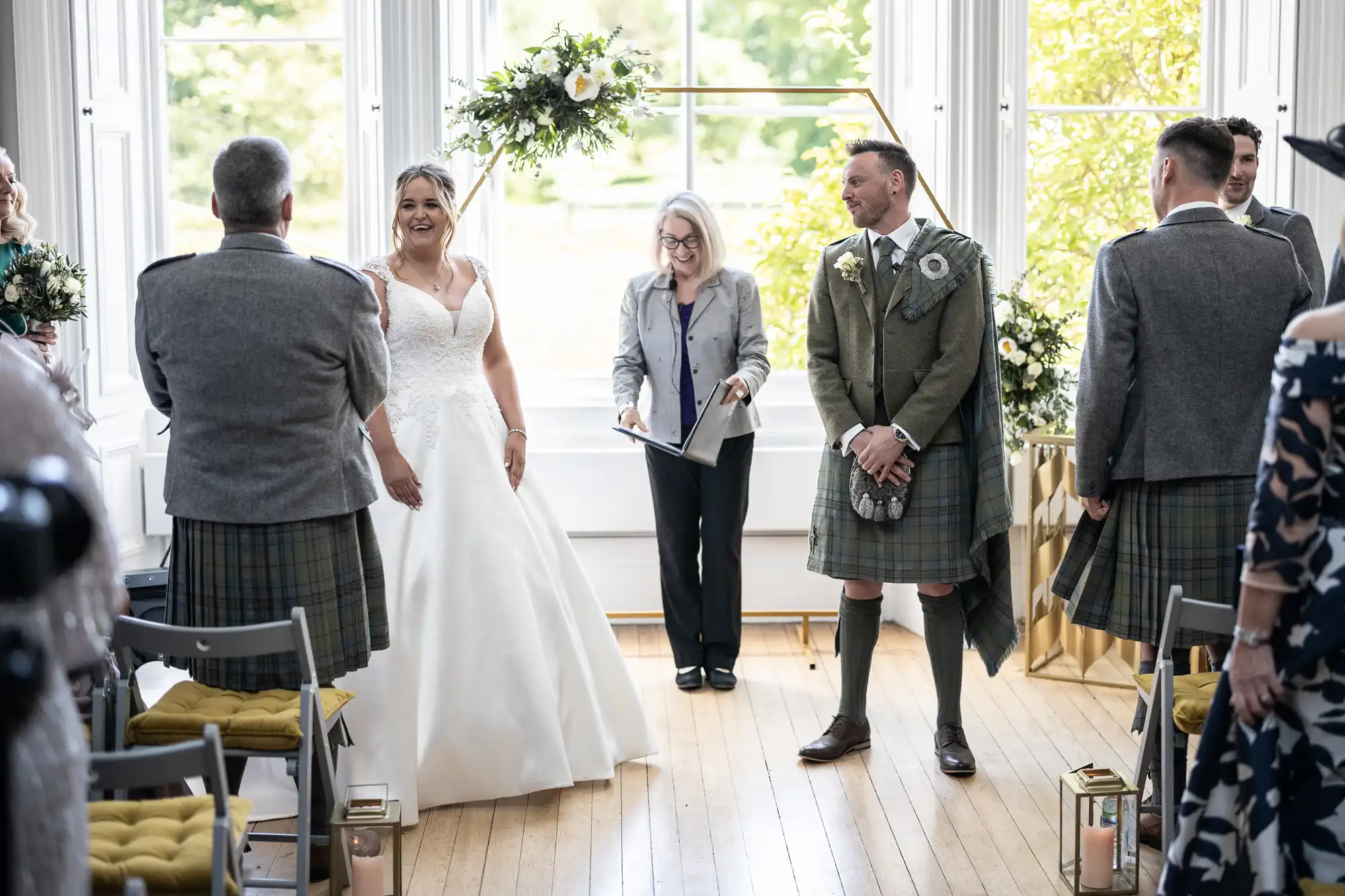 A bride and groom stand in front of an officiant during their wedding ceremony. The groom and groomsmen are wearing traditional Scottish attire, including kilts.