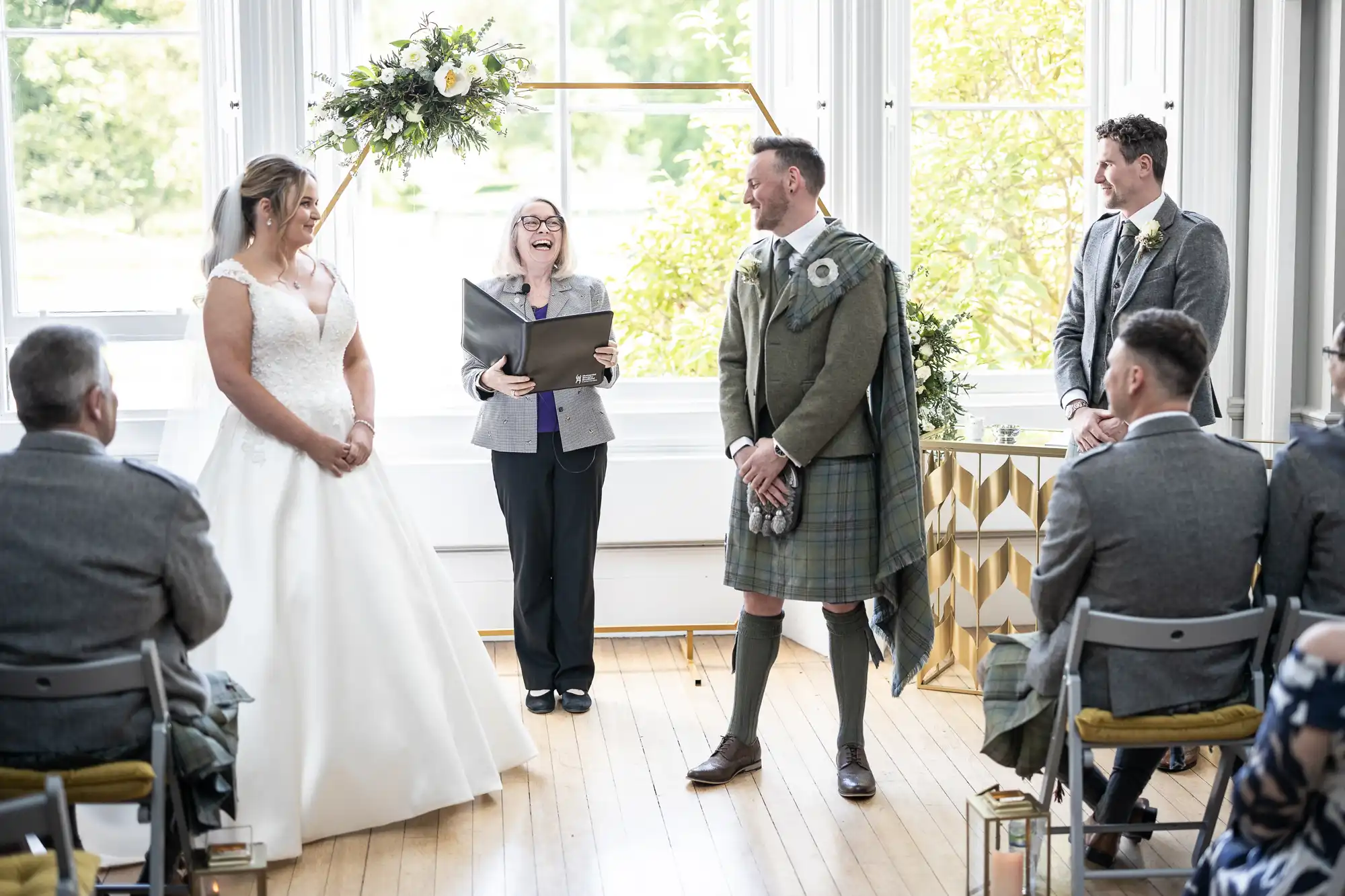 A bride and groom stand facing each other during their wedding ceremony. The officiant stands between them, smiling, while guests sit nearby. The groom is wearing a kilt.