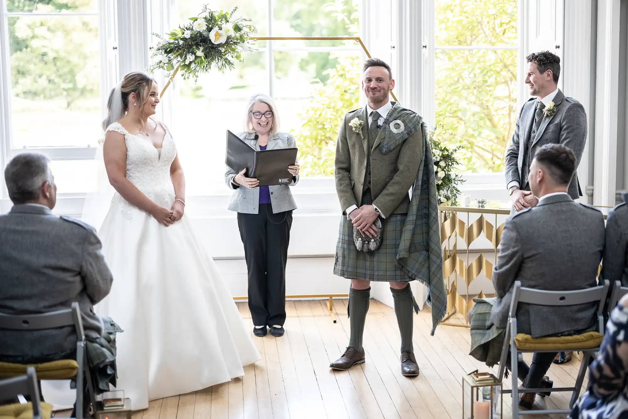 A couple stands at the altar during their wedding ceremony. The bride is wearing a white gown, and the groom is dressed in traditional Scottish attire. The officiant is reading from a book.