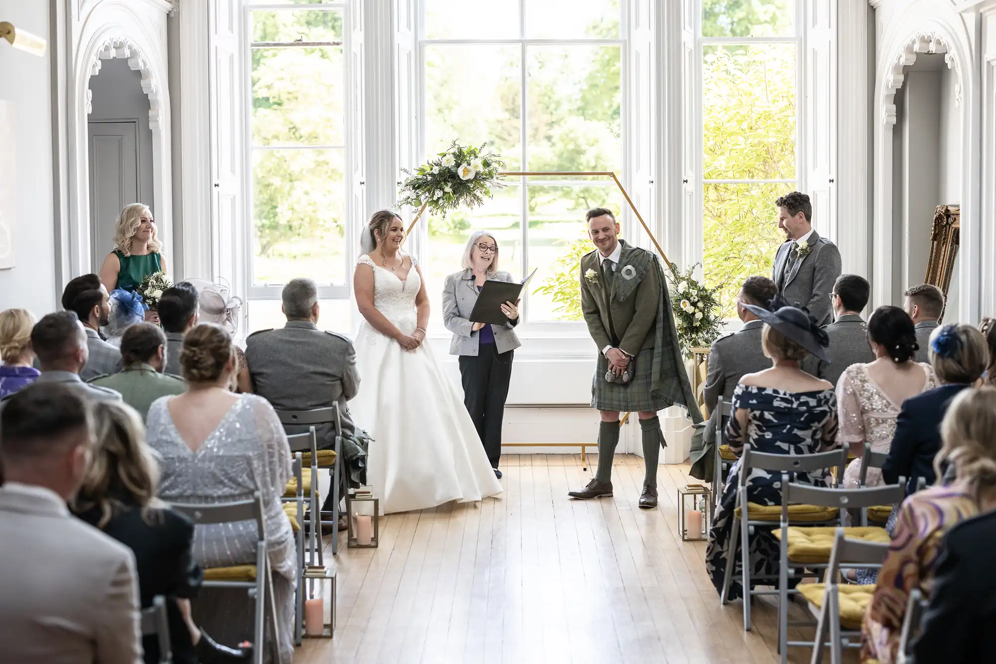 A wedding ceremony in a bright room with high windows. The bride in a white dress and the groom in a kilt stand at the front while a woman officiates. Guests are seated, watching the couple.