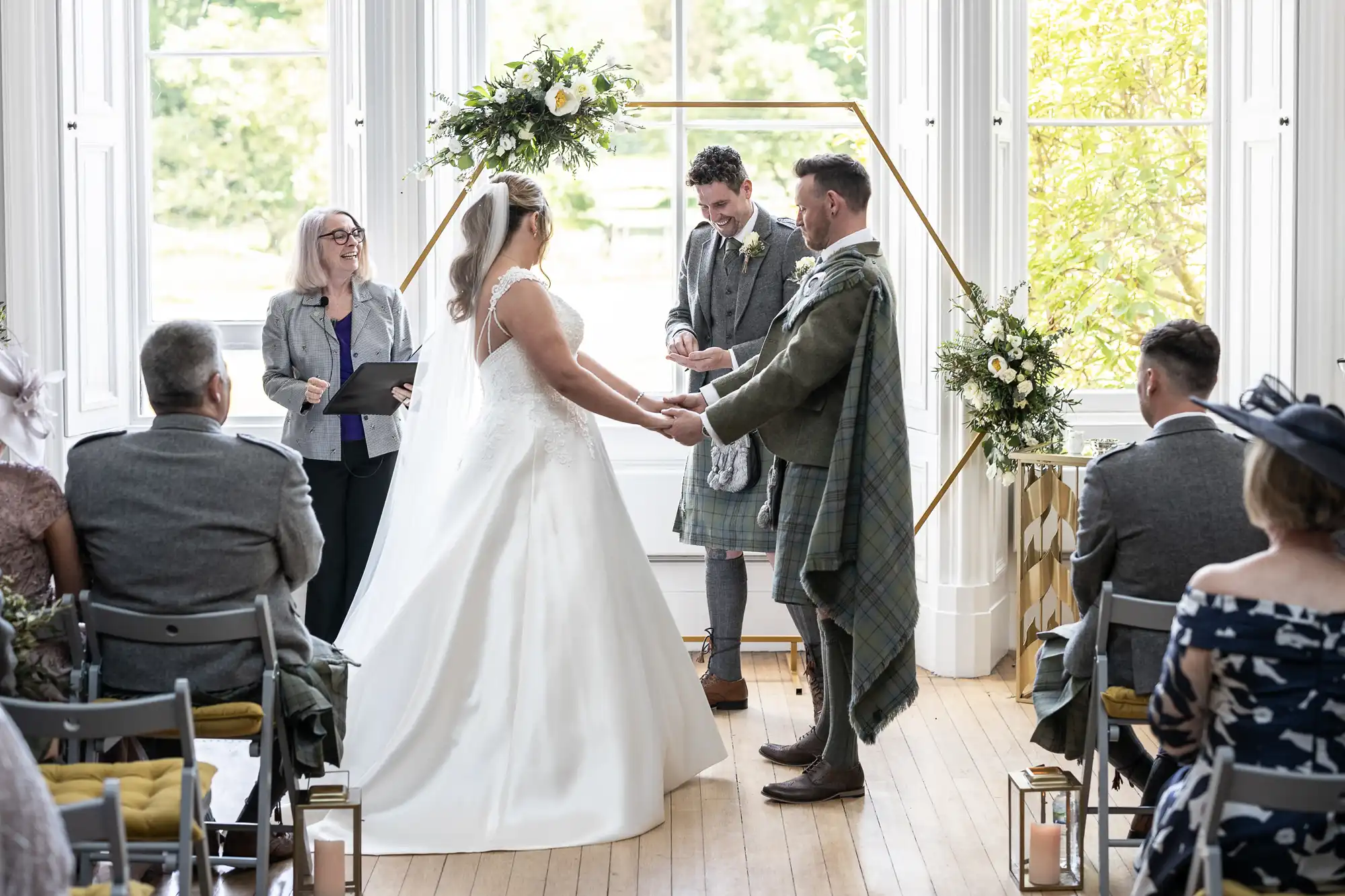 A couple stands at the altar, holding hands during a wedding ceremony. The officiant and another individual are present. Guests are seated, observing in a brightly lit room with large windows.