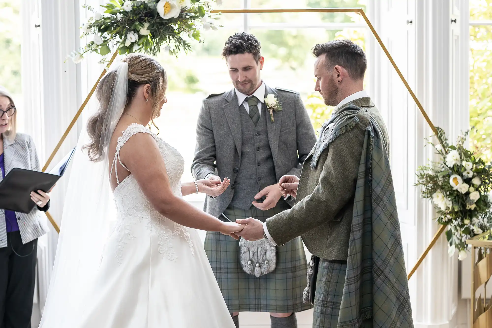 A couple in wedding attire holding hands, facing each other, in front of a person officiating the ceremony. The groom and another man wear traditional attire with kilts.