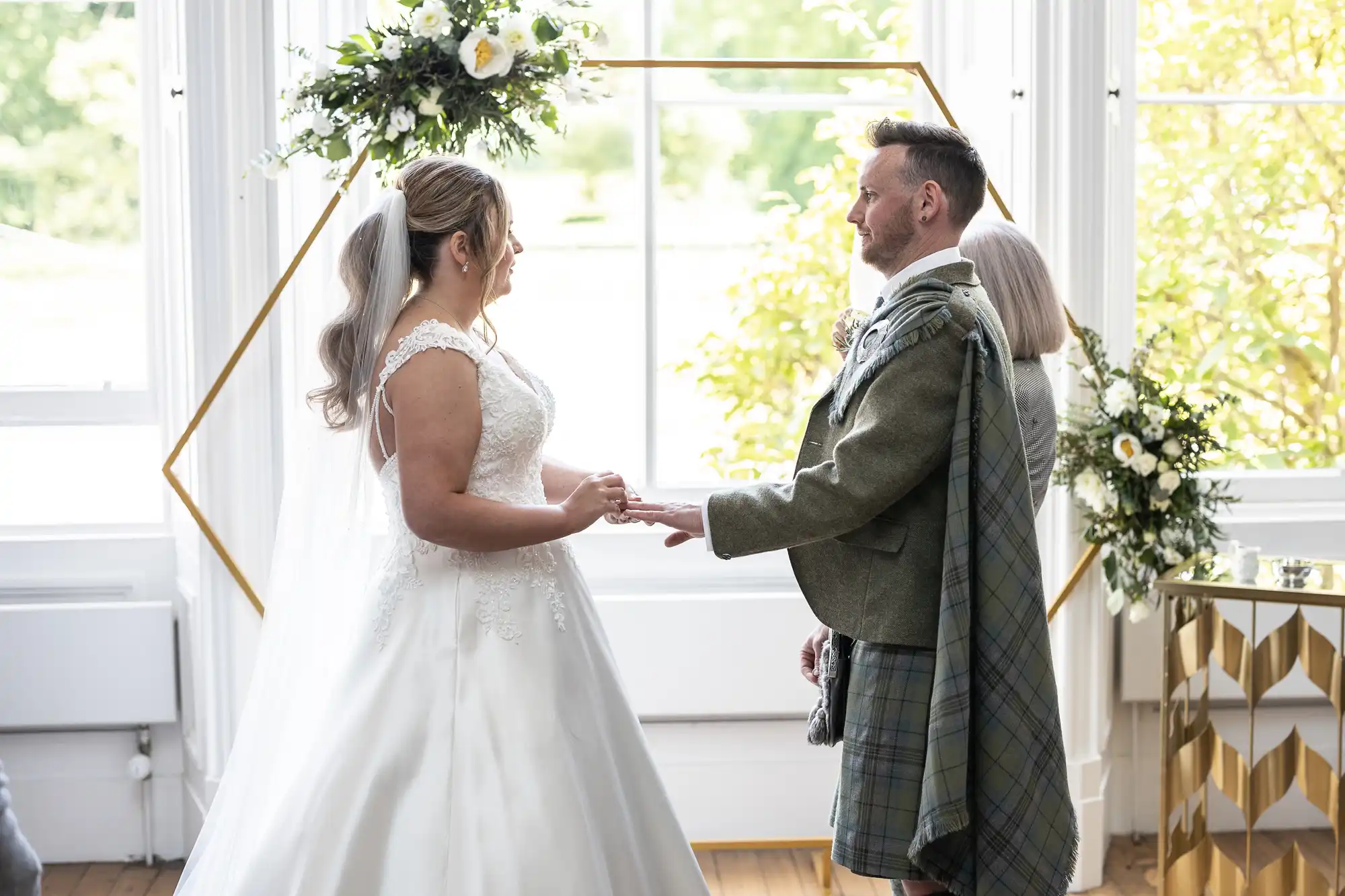 A bride and groom exchange rings in a bright room adorned with flowers and a hexagonal arch. The groom wears a traditional kilt outfit, and the bride is dressed in a white gown with a veil.