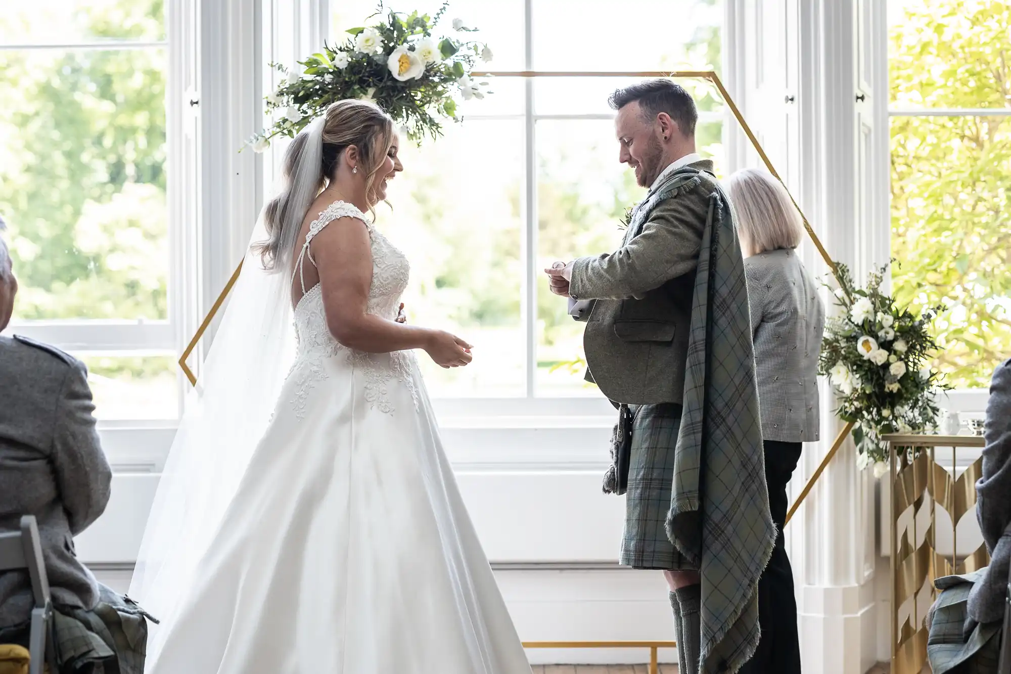 A couple stands facing each other during their wedding ceremony, with the groom in a kilt and the bride in a white gown, while an officiant stands nearby. Floral arrangements and a window are in the background.