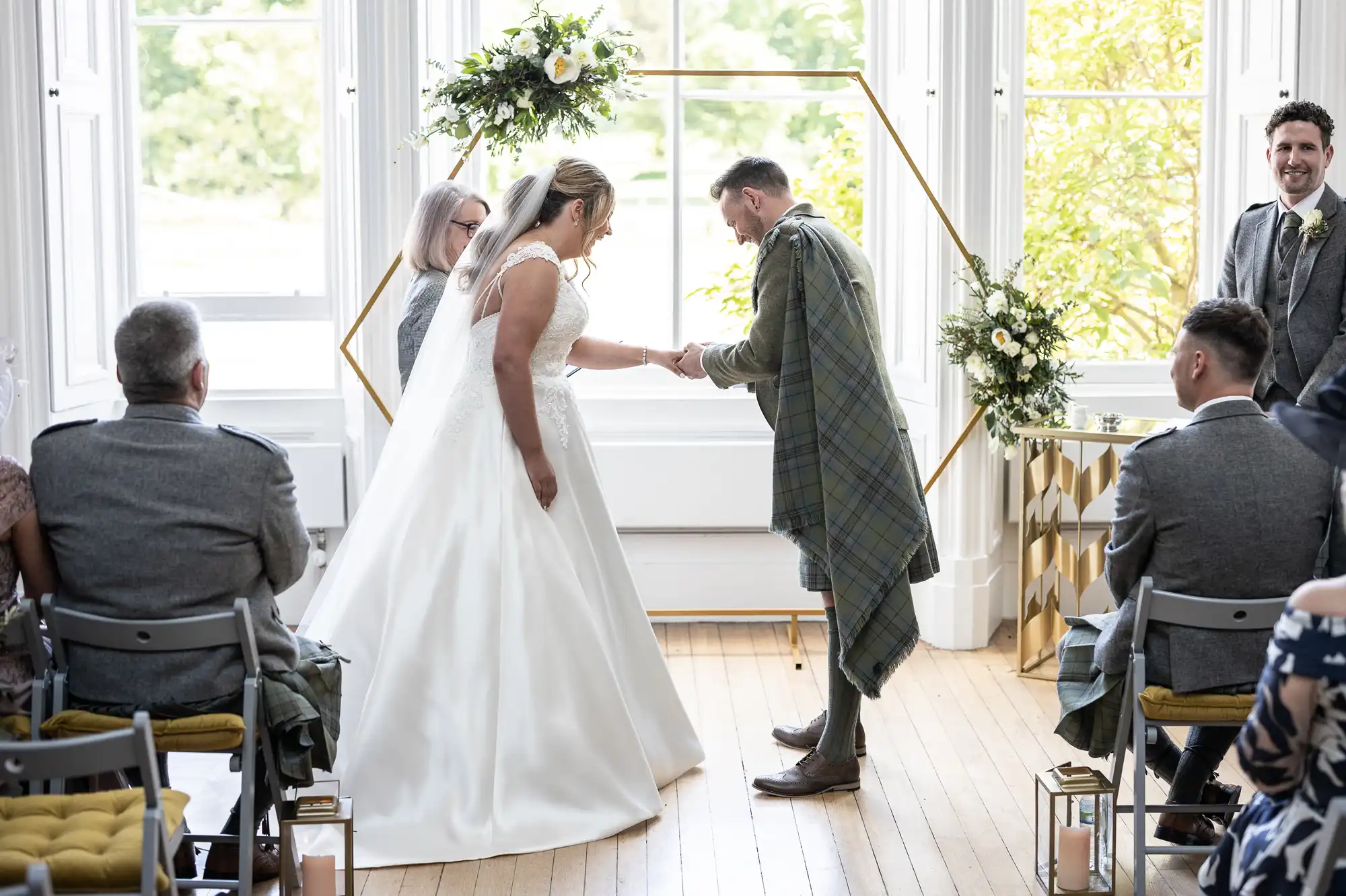 A bride and groom exchange rings during a wedding ceremony in a bright room with large windows. The groom is wearing traditional Scottish attire, including a tartan kilt. Seated guests observe.