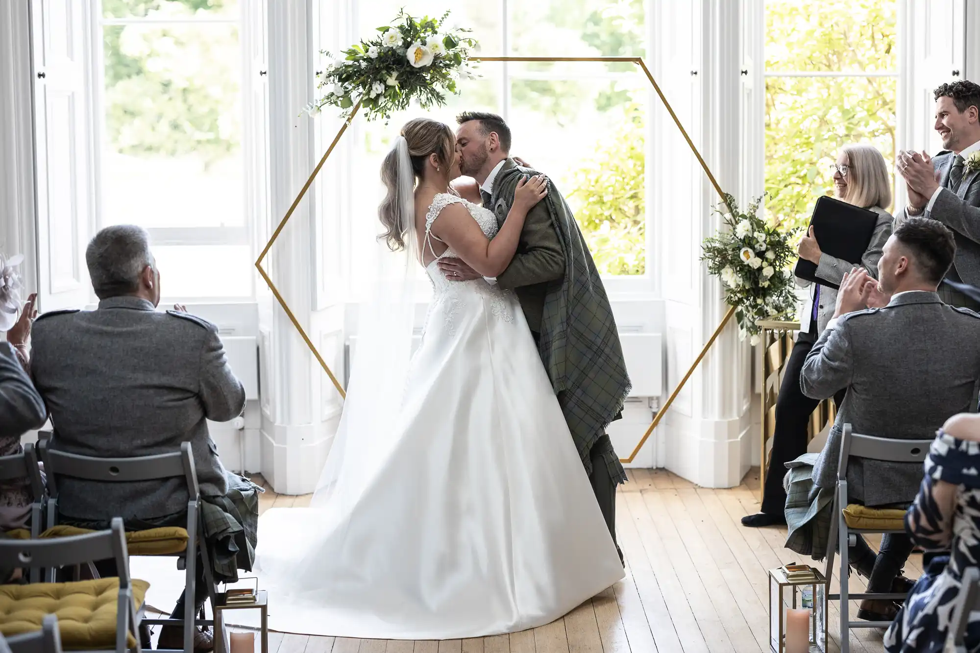 A bride and groom kiss in front of an arch during their wedding ceremony while guests sit and clap.