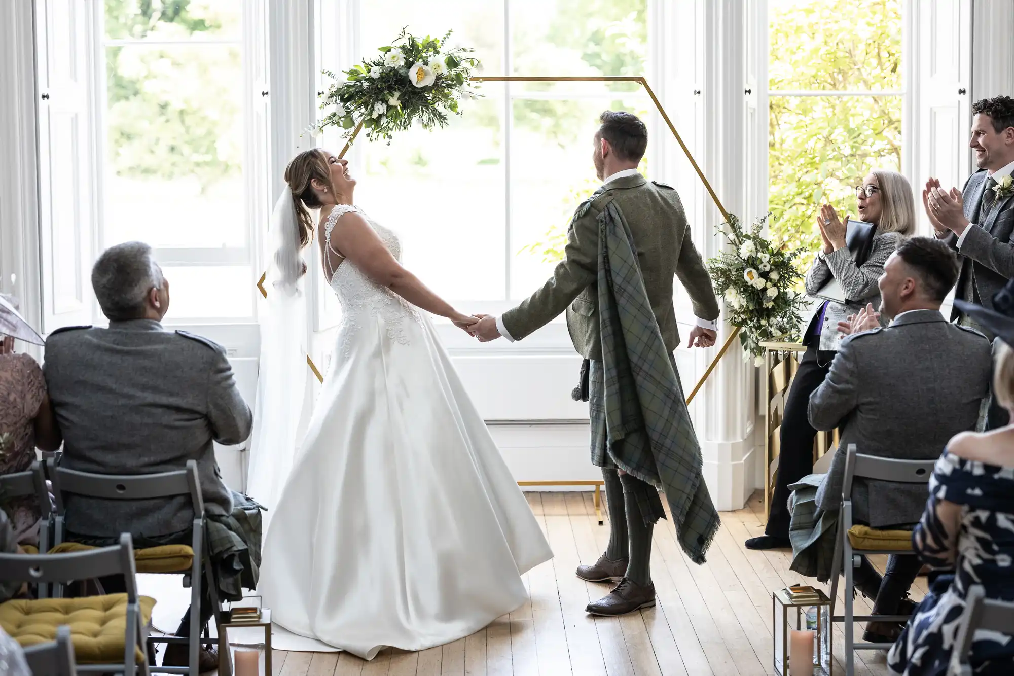 A bride and groom holding hands, celebrating at their wedding ceremony. The bride is lifting her bouquet joyfully while guests seated around them applaud. Large windows and floral decor in the background.