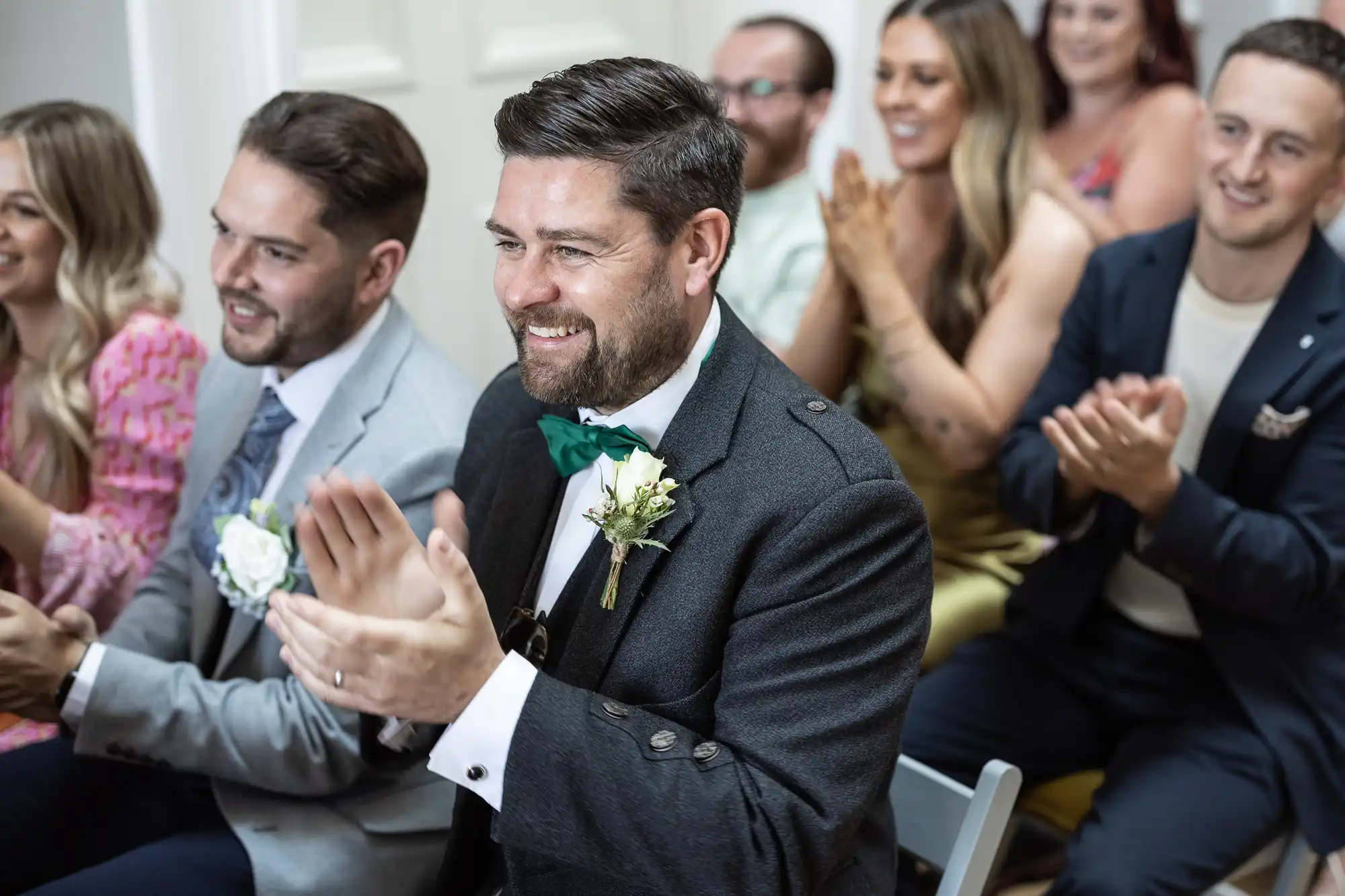 A group of people, some wearing formal attire, sit in chairs and clap their hands, seemingly pleased or entertained by an unseen event or individual.