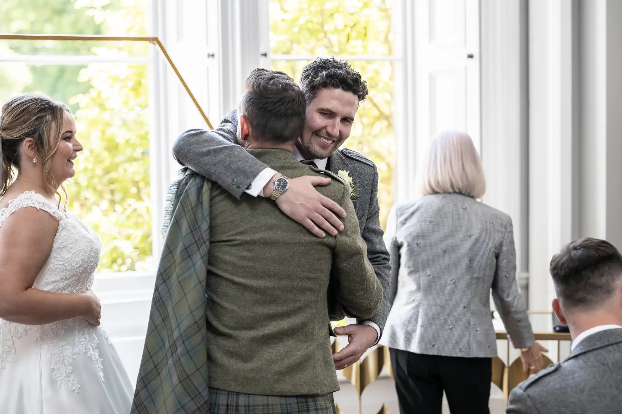 A man in a kilt and suit hugs another man while a woman in a wedding dress and an older woman in a grey jacket stand nearby in a brightly lit room.