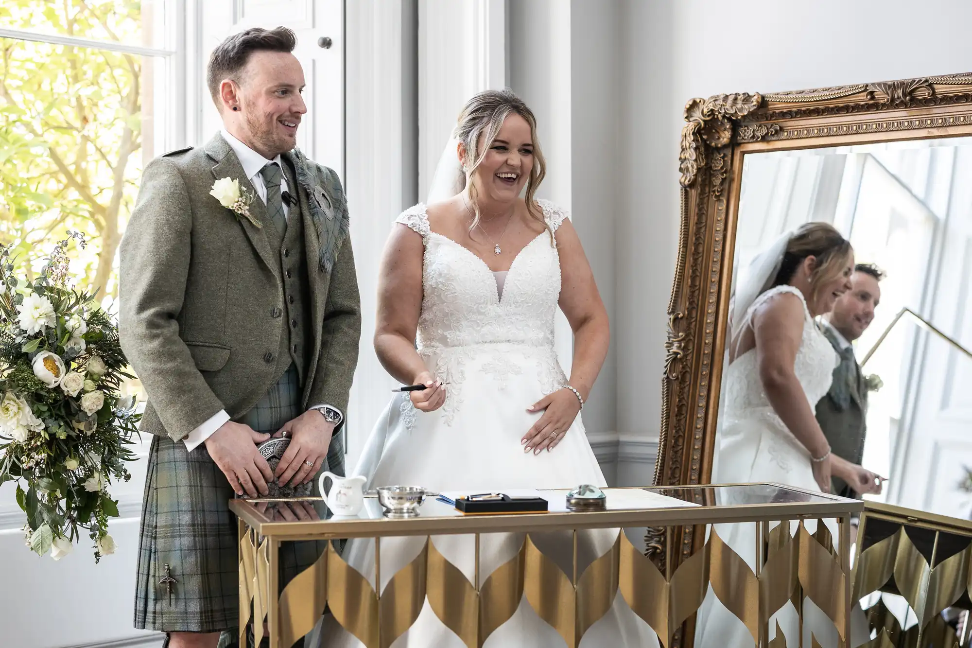 A smiling couple stands indoors in wedding attire, the groom wearing a kilt and the bride in a white dress, next to a table with various items and a large mirror reflecting their image.