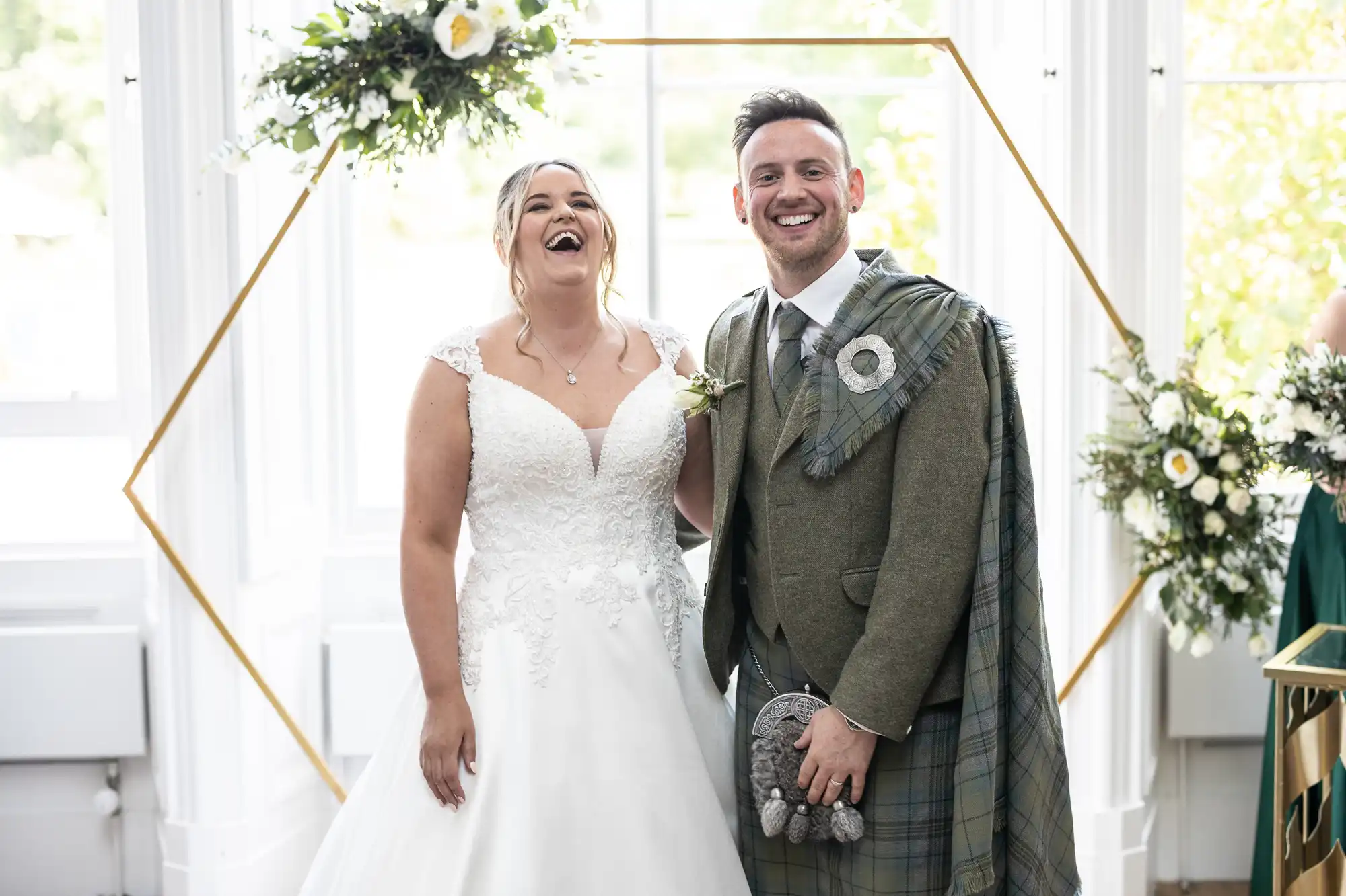 A bride and groom stand together, smiling and laughing. The bride is in a white sleeveless gown, while the groom wears a grey suit with a tartan kilt. They are in front of a floral arch indoors.