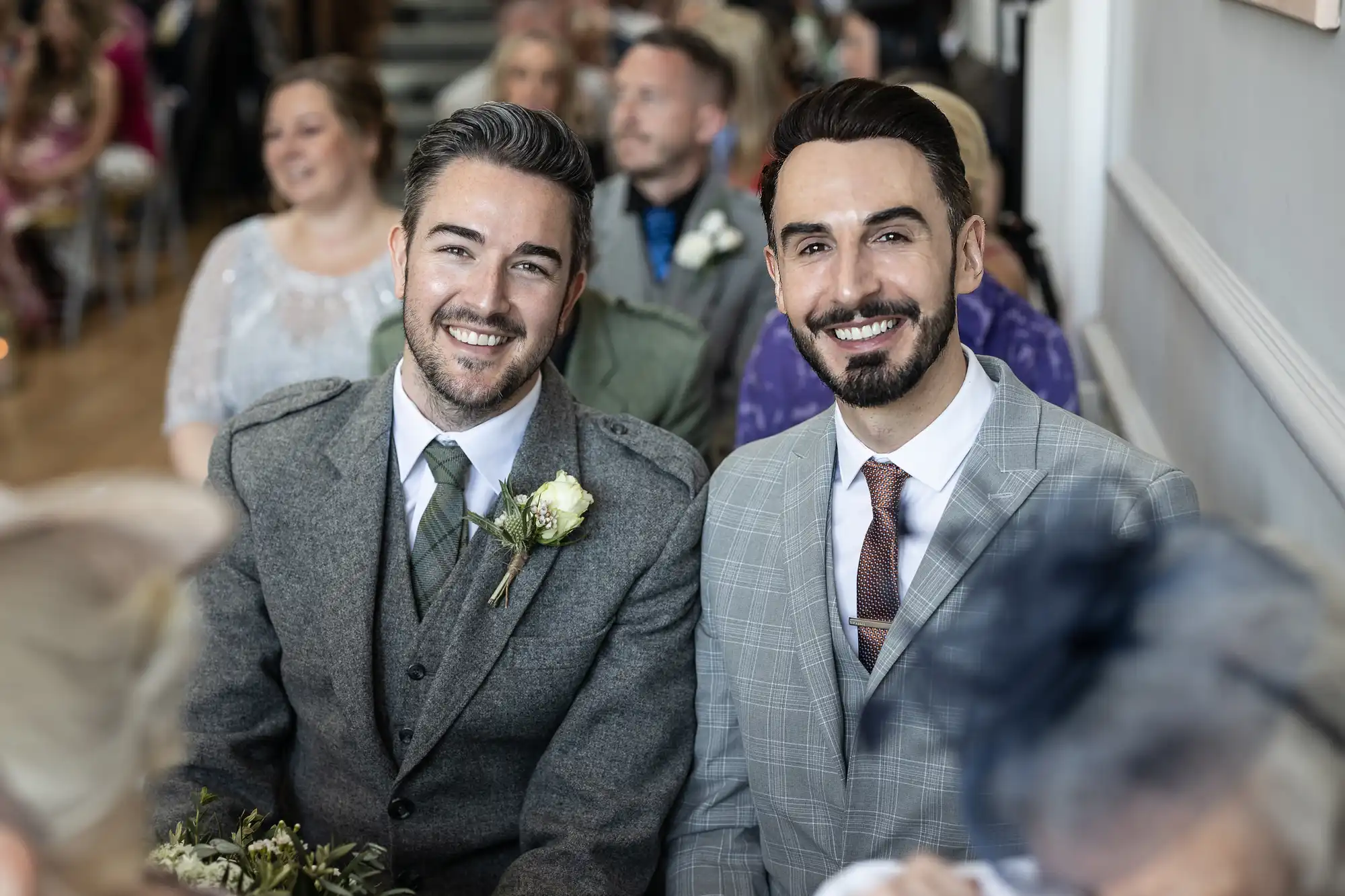 Two men wearing suits smile while seated indoors among a group of people. One man has a boutonnière pinned to his lapel.