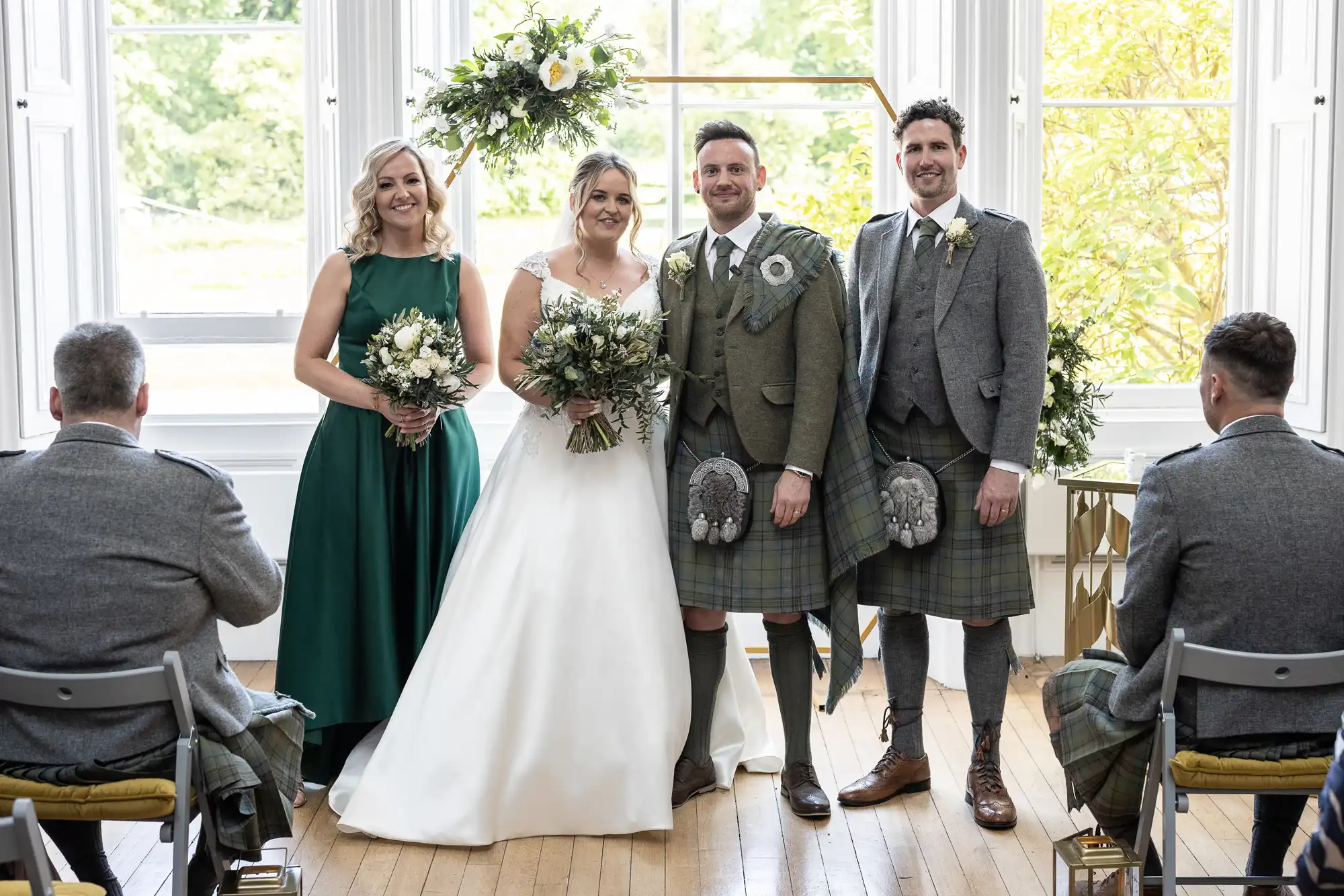 A bride in a white gown and a groom in a green kilt stand with two attendants, a woman in a green dress and a man in a green kilt, in a bright room with large windows and floral decor.