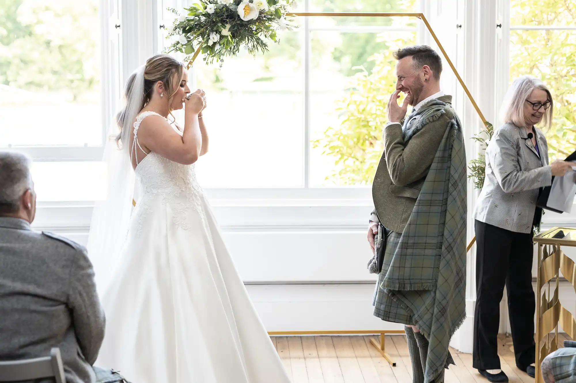 A bride in a white dress and a groom in a kilt stand in a sunlit room during their wedding ceremony. The bride is wiping her eyes while the groom smiles. A woman to the side is holding a book.