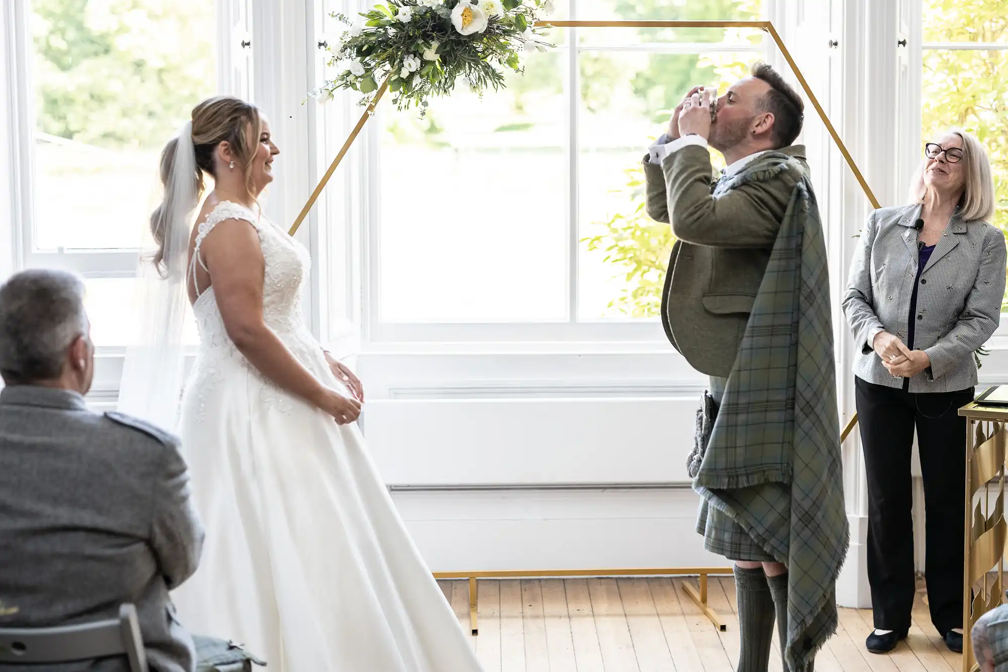 A couple stands at the altar during a wedding ceremony. The groom, dressed in a kilt, is drinking from a cup while the bride watches. An officiant and a guest are also present.