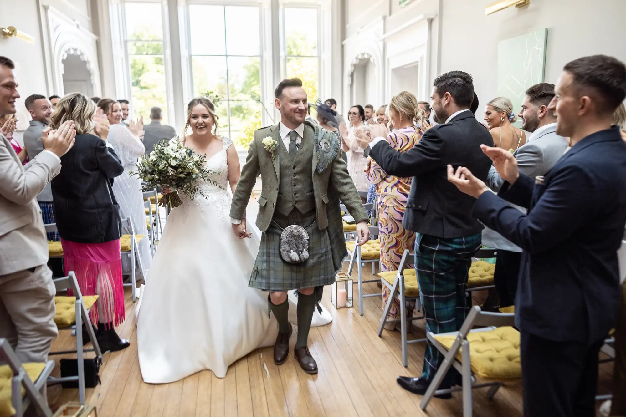 A newlywed couple walks down the aisle in a bright room as guests clap and cheer. The bride wears a white gown, and the groom wears a kilt and jacket.