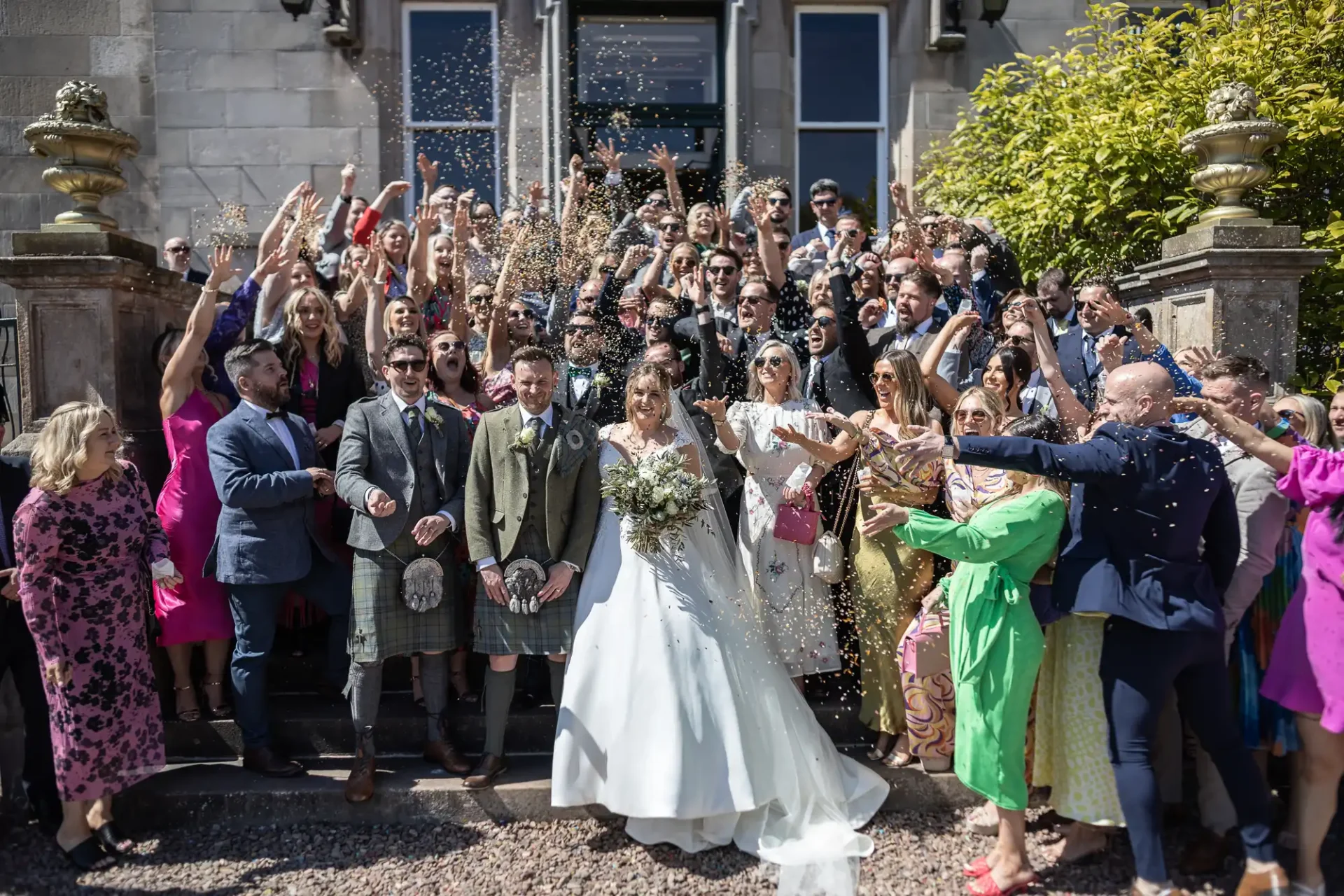 A wedding couple stands surrounded by a group of people throwing confetti outside a building. The bride is in a white dress and the groom is in traditional Scottish attire.