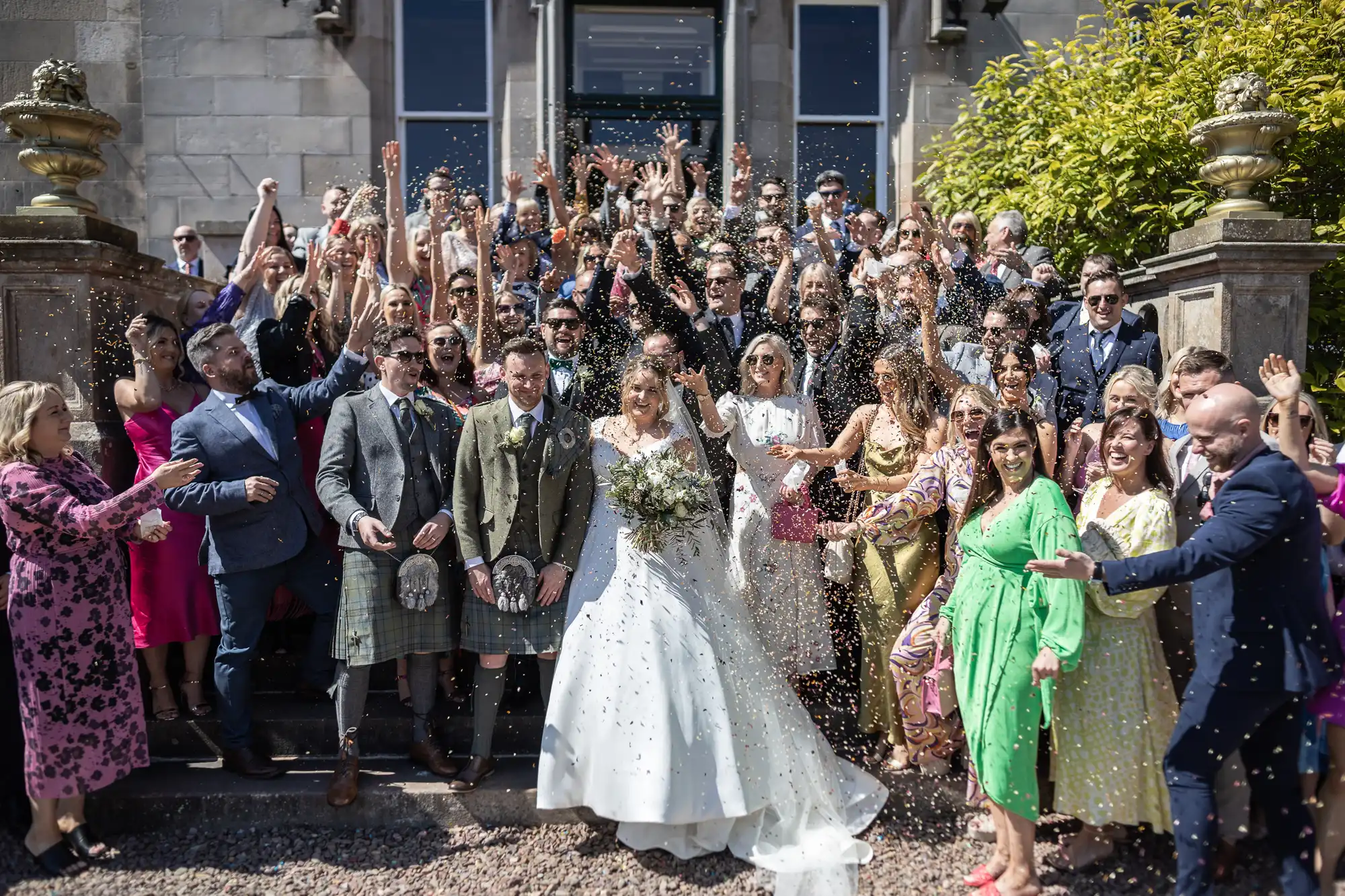 A bride and groom stand surrounded by guests outside a building. The group is celebrating, with some people tossing confetti. The sun is shining, and everyone is dressed in formal attire.