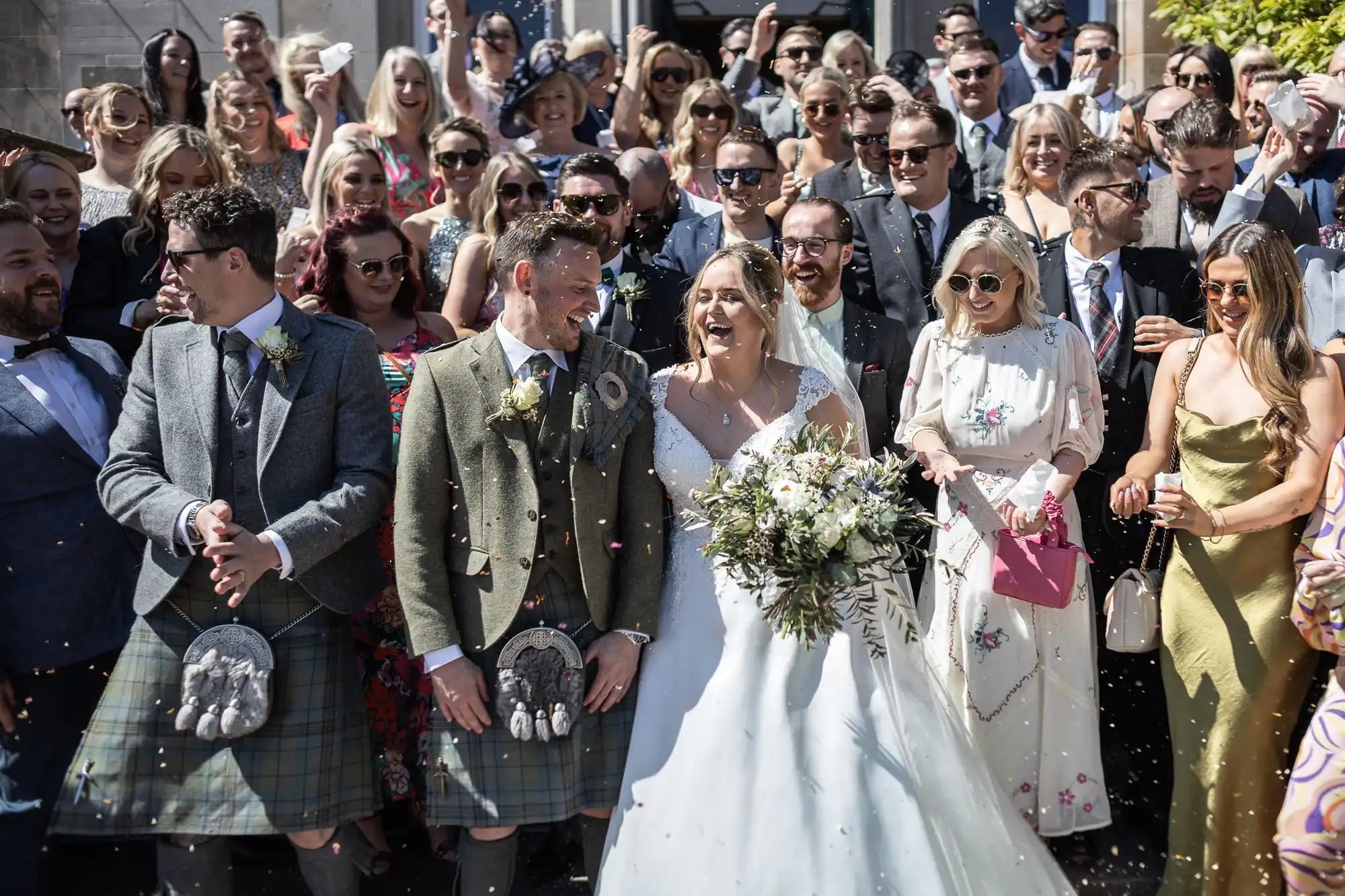 A bride and groom in wedding attire stand smiling among a large group of well-dressed guests during an outdoor celebration. Some guests are wearing sunglasses, and flower petals are in the air.