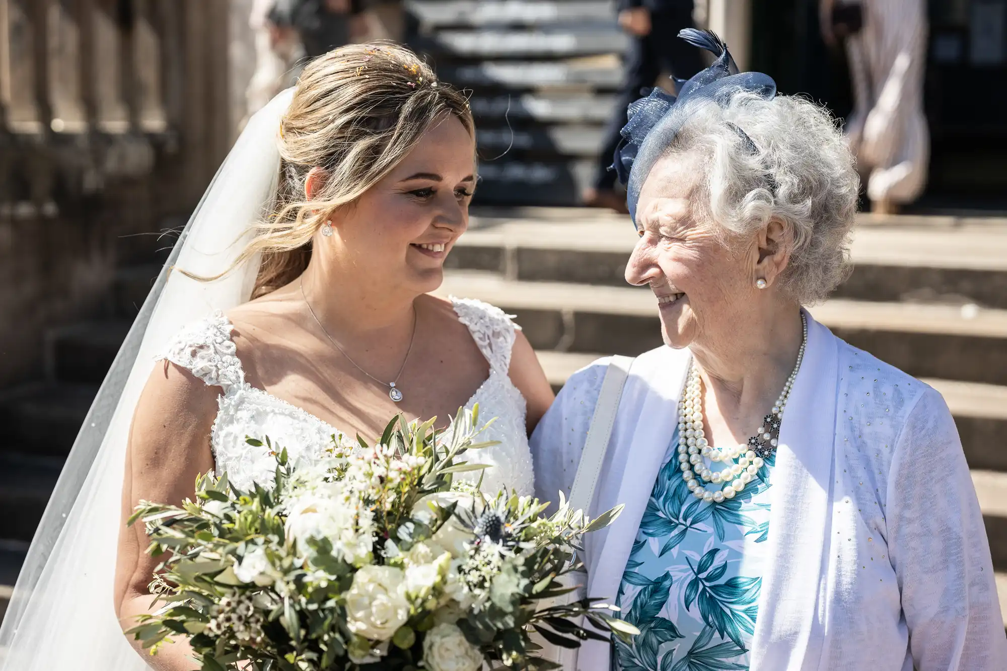 A bride in a white dress and veil smiles while holding a bouquet of flowers and standing beside an elderly woman dressed in white with a floral dress and blue fascinator. They are on outdoor steps.