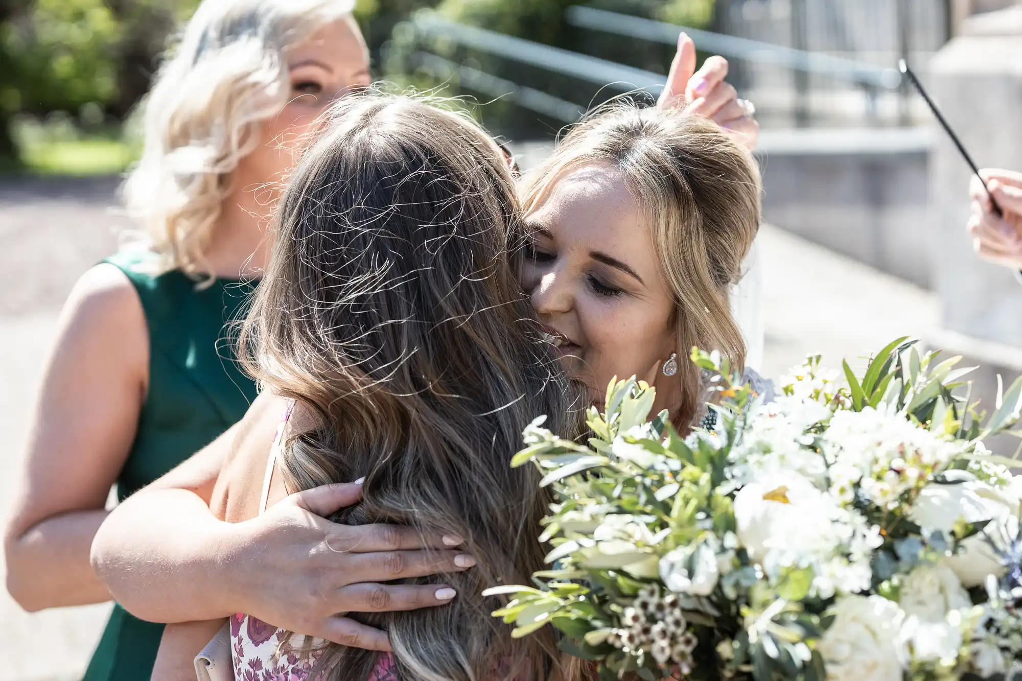 Two women embracing at an outdoor event, while one holds a bouquet of white flowers. A third person is in the background, smiling.