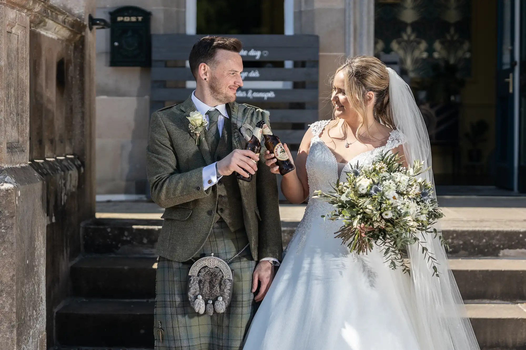 A groom in a kilt and a bride in a white dress toast with bottled drinks, standing on outdoor steps. The bride holds a bouquet, and they are exchanging smiles.