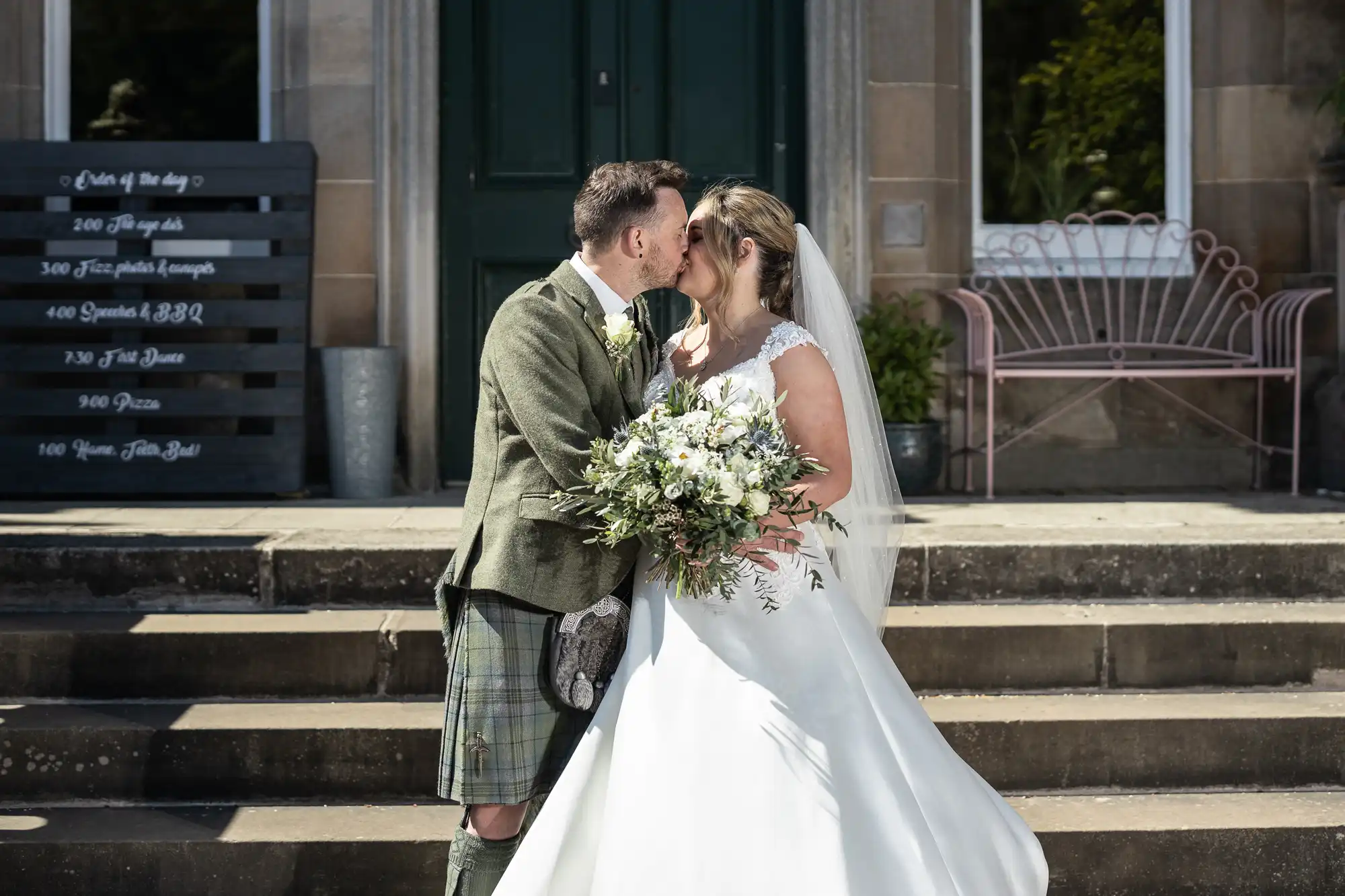 A bride and groom in traditional attire kiss on stone steps next to a wedding schedule board. The bride holds a bouquet of white flowers.